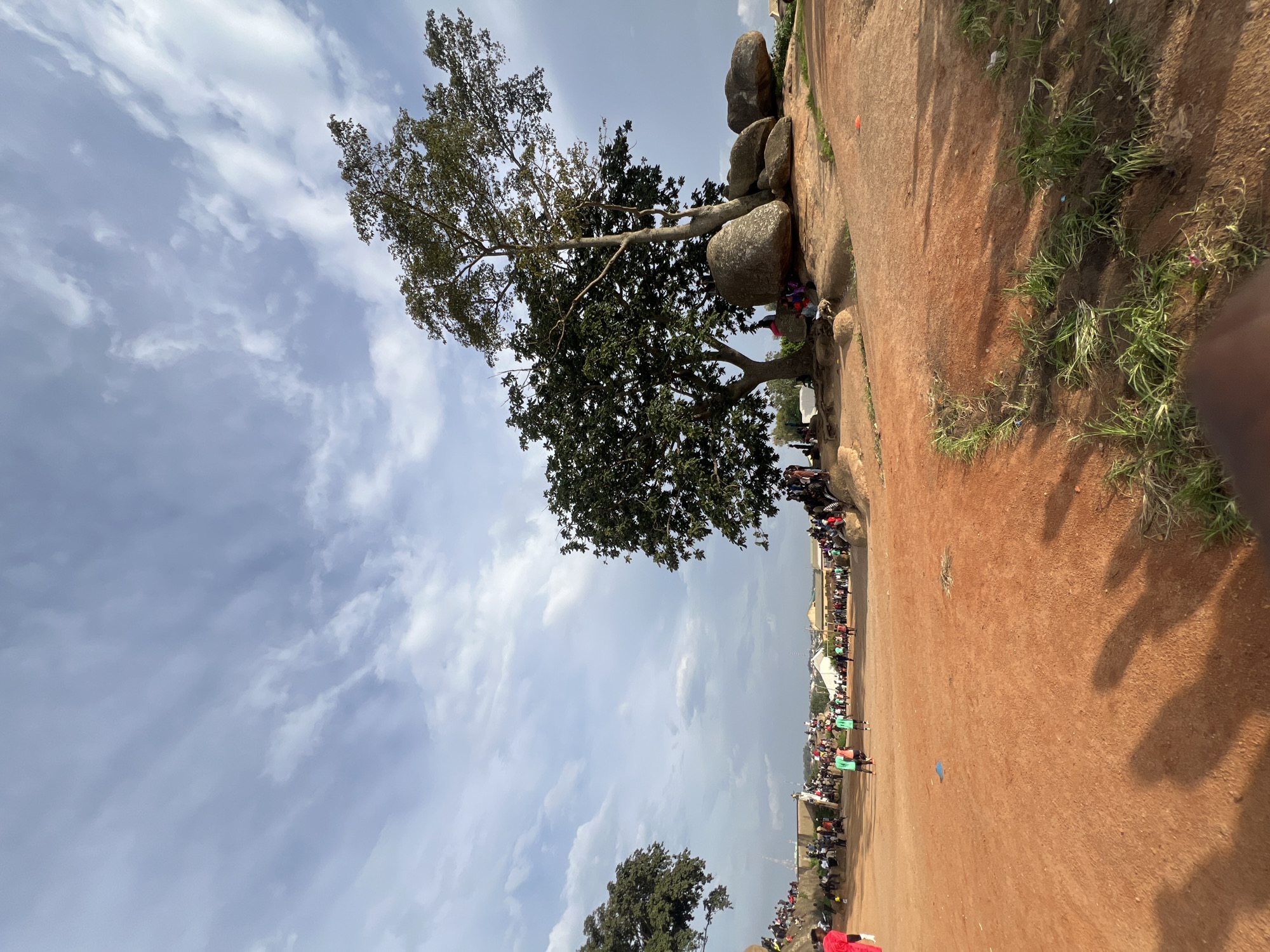 This is a vertically oriented, full-shot photograph taken outdoors in Jos, Nigeria. The scene depicts a gathering of people on a reddish-brown, dusty ground, with large boulders and a prominent tree to the left. The sky is cloudy, suggesting daytime with potentially overcast conditions.
The subjects are a large crowd of people, many of whom appear to be standing or walking along a path or open area. They are dressed in casual clothing, with some individuals wearing green shirts, possibly indicating a team or group affiliation. The large tree with its dense foliage and spreading branches is a central element of the landscape, with several large, smooth boulders nestled around its base and extending up a rocky incline.
The activity taking place appears to be a gathering or event, possibly a festival, market, or community assembly, given the large number of people. Some individuals are positioned on the rocky area, observing the scene below. There are also structures and buildings visible in the distance, suggesting a semi-urban or rural setting on the periphery of a town.
Notable details include the vibrant red earth, the texture of the boulders, and the contrast between the natural landscape and the human presence. The lighting suggests it is likely midday, but the clouds diffuse the sunlight. No text is visible in the image. The overall mood appears to be one of communal activity and a lively atmosphere.