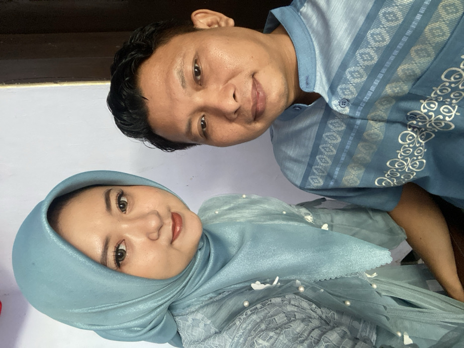 This image depicts two adult individuals, a female and a male, posing for a close-up portrait indoors in Tambakrejo, Indonesia. The female, positioned on the left and slightly closer to the viewer, wears a light blue hijab covering her head and neck, along with a matching garment featuring lace detailing, white pearls, and small white floral appliques. She has light-colored eyes, possibly contact lenses, and visible makeup including eyeliner and lipstick. The male, situated to the right and slightly behind the female, wears a short-sleeved blue collared shirt with a mandarin collar. His shirt displays horizontal striped patterns in varying shades of blue with geometric designs, and white ornamental patterns on the lower visible section. Both individuals are looking directly at the camera with neutral expressions, appearing to offer slight smiles. The background consists of a plain, light-colored wall, with a darker vertical element partially visible on the far right. The scene is illuminated by consistent, even indoor lighting.