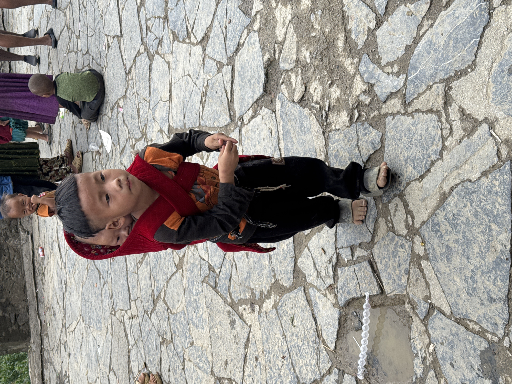 In Yên Minh Commune, Vietnam, a child wearing black pants, a black and orange long-sleeve shirt, and sandals stands on a stone pathway. A red carrier is wrapped around the child, holding a baby. In the background, there are glimpses of other people, including adults and children. The pathway is composed of large, irregularly shaped stones. A string of white objects lies near a small pool of water on the stone pathway.