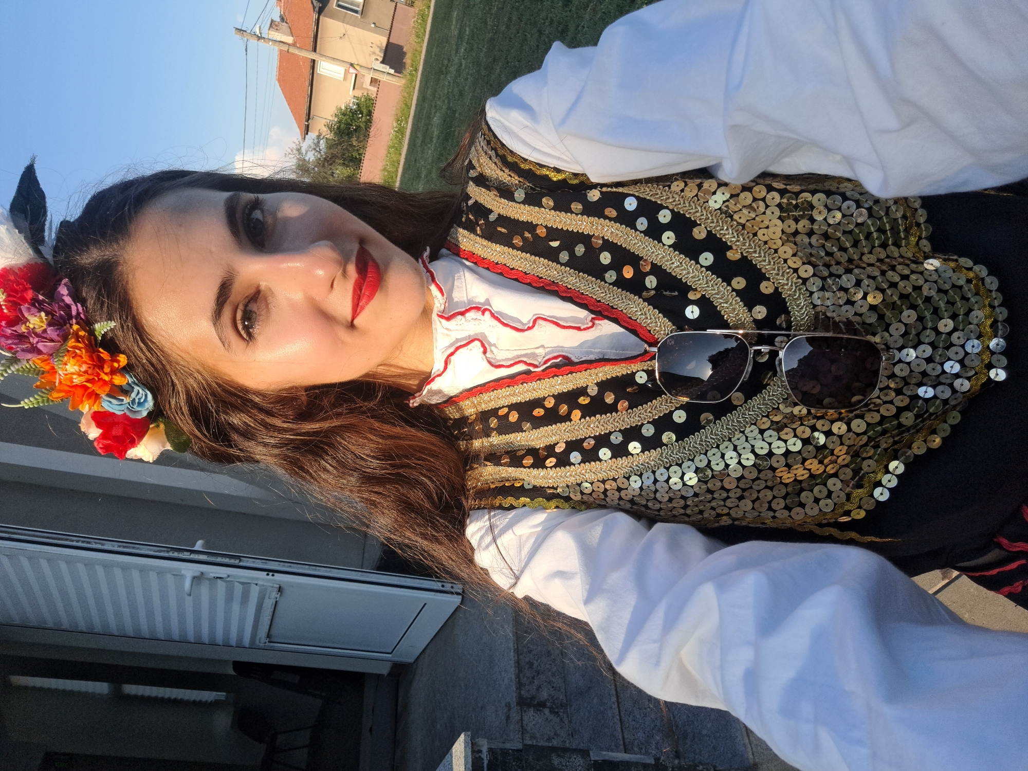 A young woman with long, dark hair adorned with a colorful flower crown poses for a selfie outdoors. She is wearing a traditional, richly decorated outfit consisting of a white ruffled blouse and a black vest embellished with gold sequins and trim. Sunglasses are perched on her vest. The background shows a residential building with a red-tiled roof, green grass, and a clear blue sky, suggesting it is daytime. The warm lighting indicates it might be late afternoon or early evening. The woman has bright makeup, with red lipstick and mascara, and she is smiling, conveying a cheerful mood. The overall scene appears festive and celebratory.