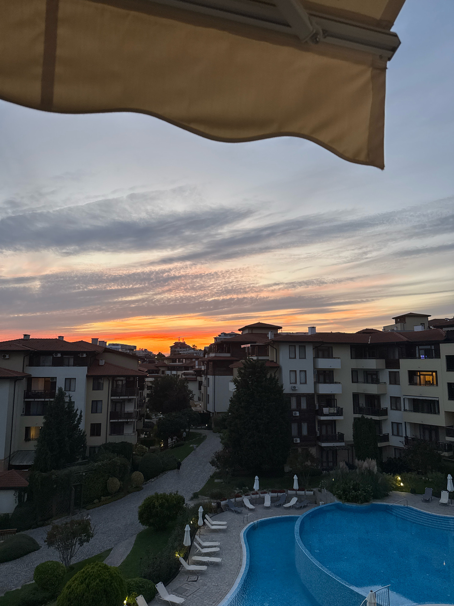 The image shows a twilight view over a resort complex in Sveti Vlas, Bulgaria. The sky displays a vibrant sunset with streaks of orange and pink clouds against a pale blue backdrop. In the foreground, a beige awning partially obscures the top of the frame. Below the sky, a cluster of multi-story buildings with terracotta roofs and balconies is visible. These buildings appear to be part of the resort, with windows indicating occupancy. A winding path leads through landscaped grounds with trees and bushes. To the right, a blue swimming pool with lounge chairs and umbrellas is situated. The lighting suggests it is late evening, with some lights illuminated in the buildings and along the pathways. No people are clearly discernible in the image, though the setting implies a residential or vacation area.