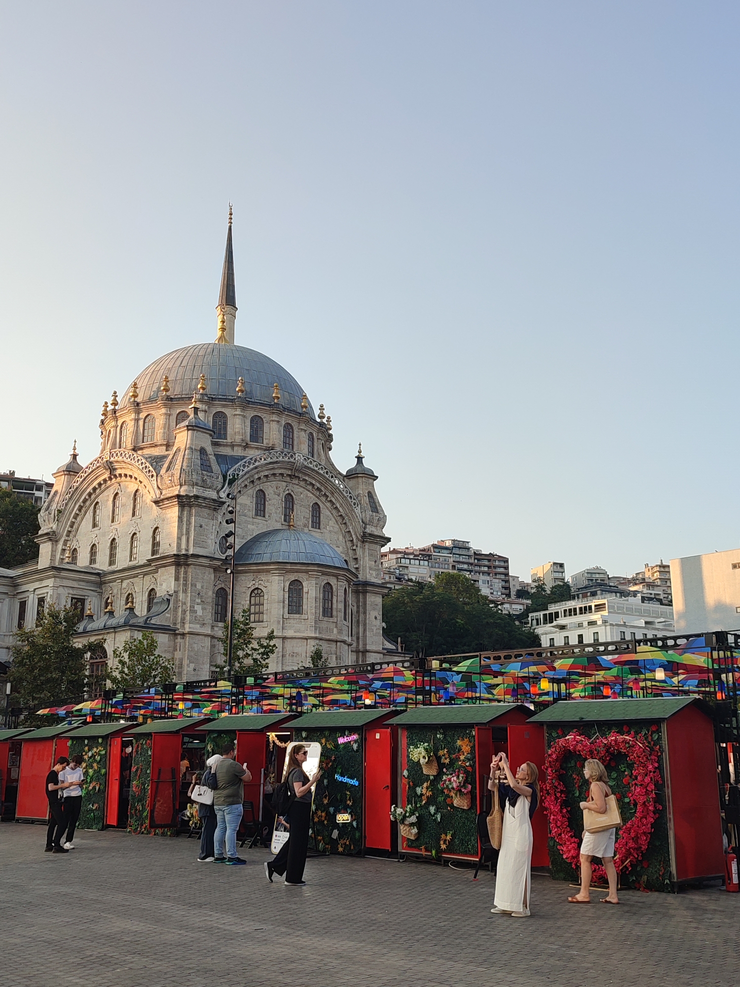 The image depicts a street scene in Beyoğlu, Turkey, with a prominent mosque in the background and several market stalls in the foreground. The mosque, a grand stone structure with a large dome and a tall minaret, is bathed in the warm light of what appears to be late afternoon or early morning. In front of the mosque, a row of red market stalls with green roofs are set up. These stalls are decorated with floral arrangements, including a large heart-shaped wreath made of red flowers. Above the stalls, a colorful canopy of umbrellas provides shade.
Several people are visible in the scene. On the left, two young men are standing together, one of whom is looking at his phone. Further to the right, a woman in a black dress is posing for a photo. Next to her, a woman in a white dress is taking a picture with her phone, while another woman in a white dress and shorts stands beside a decorated stall, holding a shopping bag. The ground is paved with cobblestones. In the background, beyond the mosque, are buildings characteristic of a city on a hill, with multiple stories and balconies. The sky is clear and pale blue, suggesting fair weather. The overall atmosphere is one of a vibrant urban setting, possibly a tourist area or a lively public square.