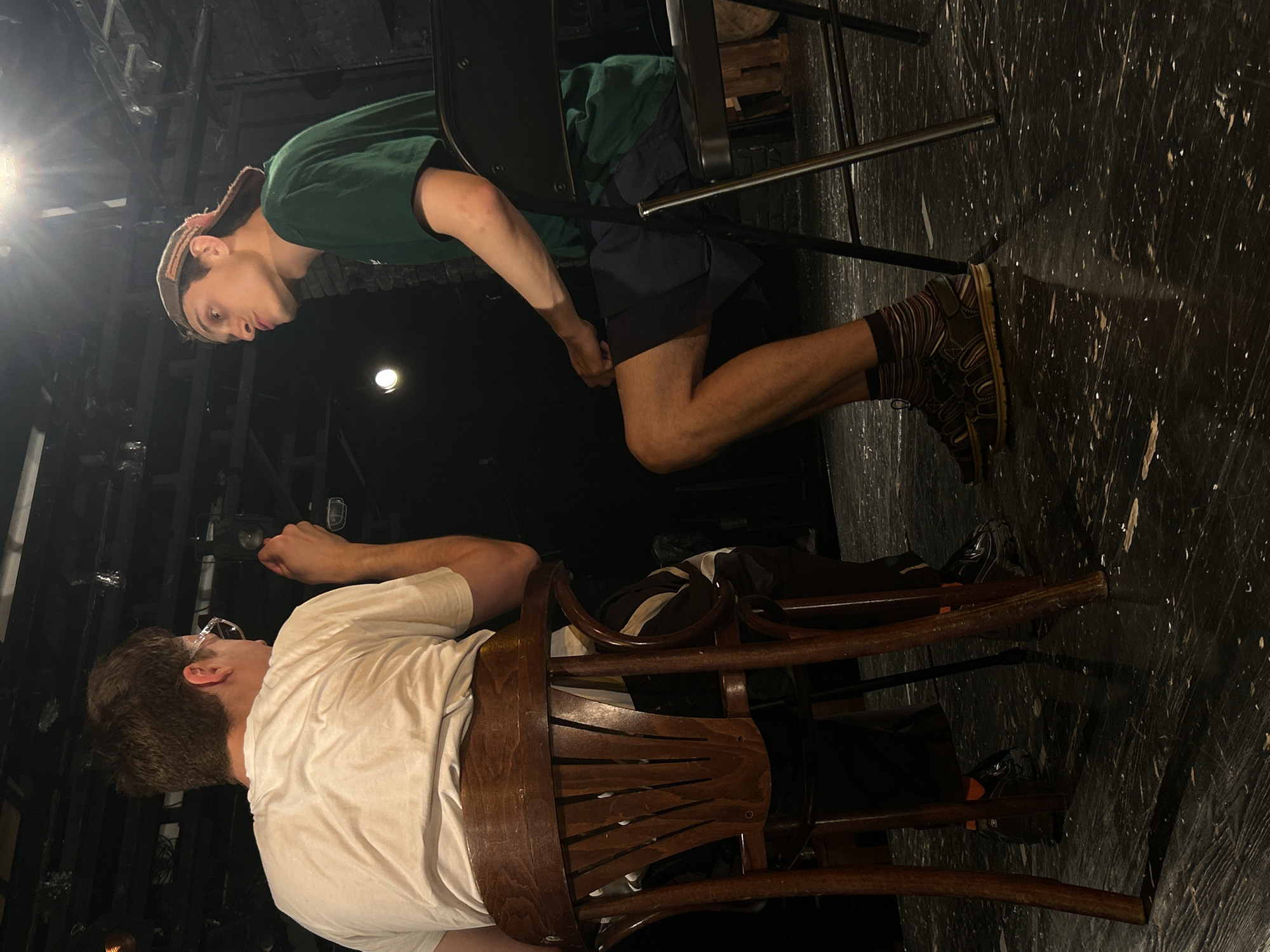 Two young men are seated on chairs on a dark, reflective stage floor. The man on the left, viewed from the back and partially from the side, wears a white t-shirt, dark pants with a stripe detail, and glasses. He is holding a cigarette or a similar object in his right hand. The man on the right, facing the other, wears a green t-shirt, dark shorts, striped socks, and sandals, with a baseball cap on his head. Both men appear to be engaged in conversation, with the man on the right looking directly at the man on the left. The environment suggests a theater or performance space, with exposed lighting fixtures and dark theatrical backdrops visible above and behind the subjects. The lighting is dramatic, with a bright overhead source casting strong highlights and shadows.