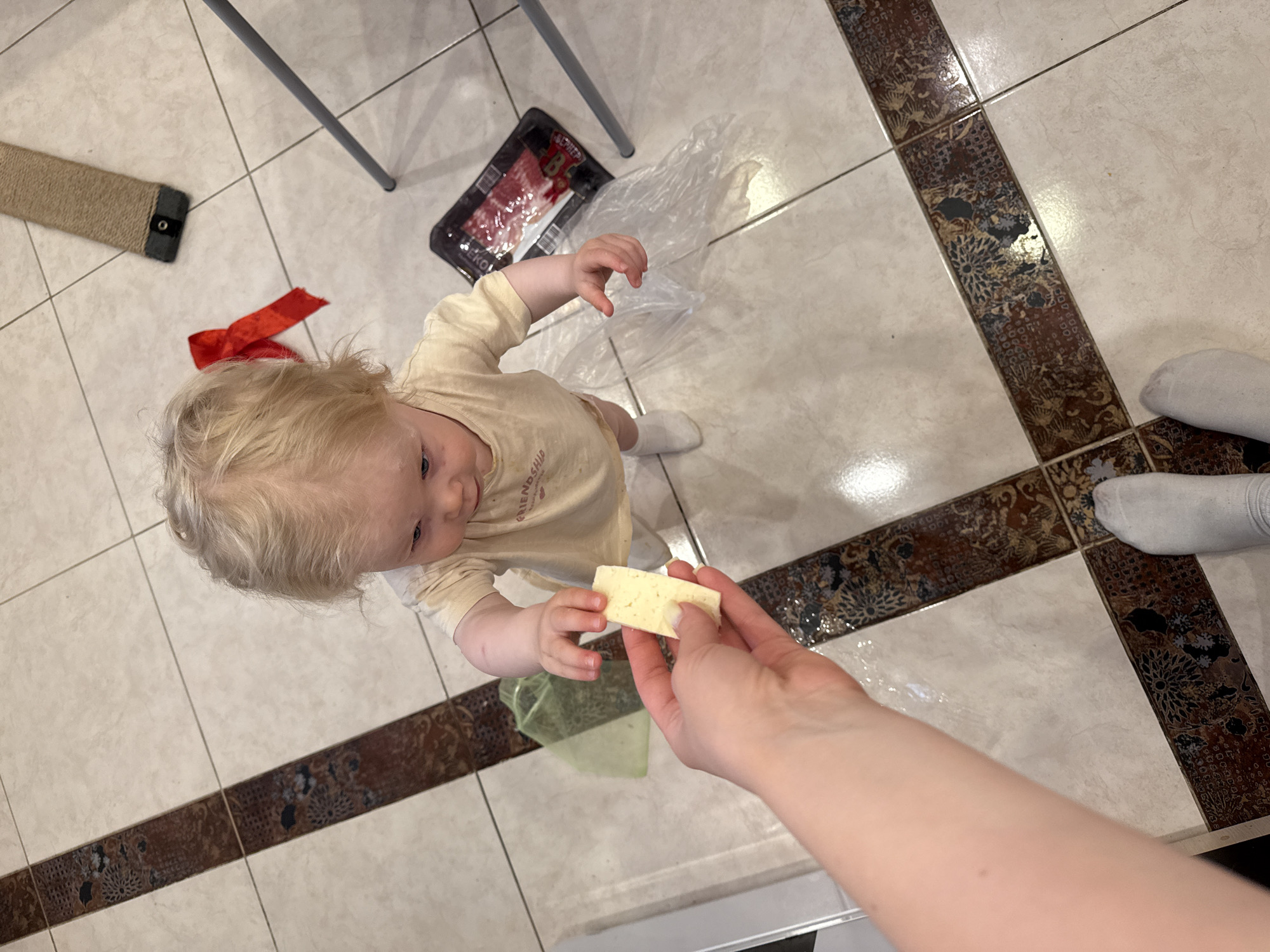 A high-angle shot shows a young child with blonde hair looking up at a piece of cheese held out by an adult's hand. The child is wearing a light-colored shirt and white socks, standing on a tiled floor with decorative brown tile borders. A package of meat, a red ribbon, a cat scratching post, a plastic bag, and legs of a table are visible in the background. The person holding the cheese has fair skin and is wearing a white sock. The location is the city of Vladimir, Russia.