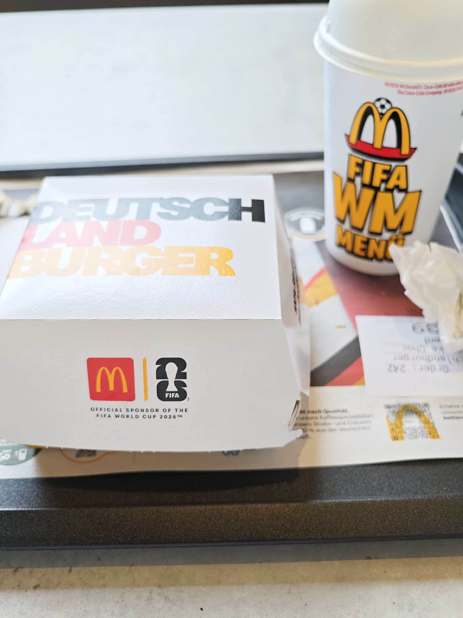 The image shows a McDonald's "Deutschland Burger" box and a promotional cup, suggesting a tie-in with a FIFA World Cup event. The box has "DEUTSCHLAND BURGER" printed on it in large, colorful letters, with "DEUTSCH" in gray and "LAND BURGER" in red and yellow, echoing the German flag colors. The cup features the McDonald's logo and text related to "FIFA WM" (World Cup) and "MENÜ" (Menu). Below the McDonald's and FIFA logos on the box, it states "OFFICIAL SPONSOR OF THE FIFA WORLD CUP 2026™". The setting appears to be a McDonald's restaurant, possibly in Tuttlingen, Germany, given the location context. The overall impression is a promotional display for a limited-edition menu item related to a sporting event. There are no people visible in the image. The lighting suggests it could be daytime, and the setting is indoors. Other visible text on a receipt includes "Order: 242".