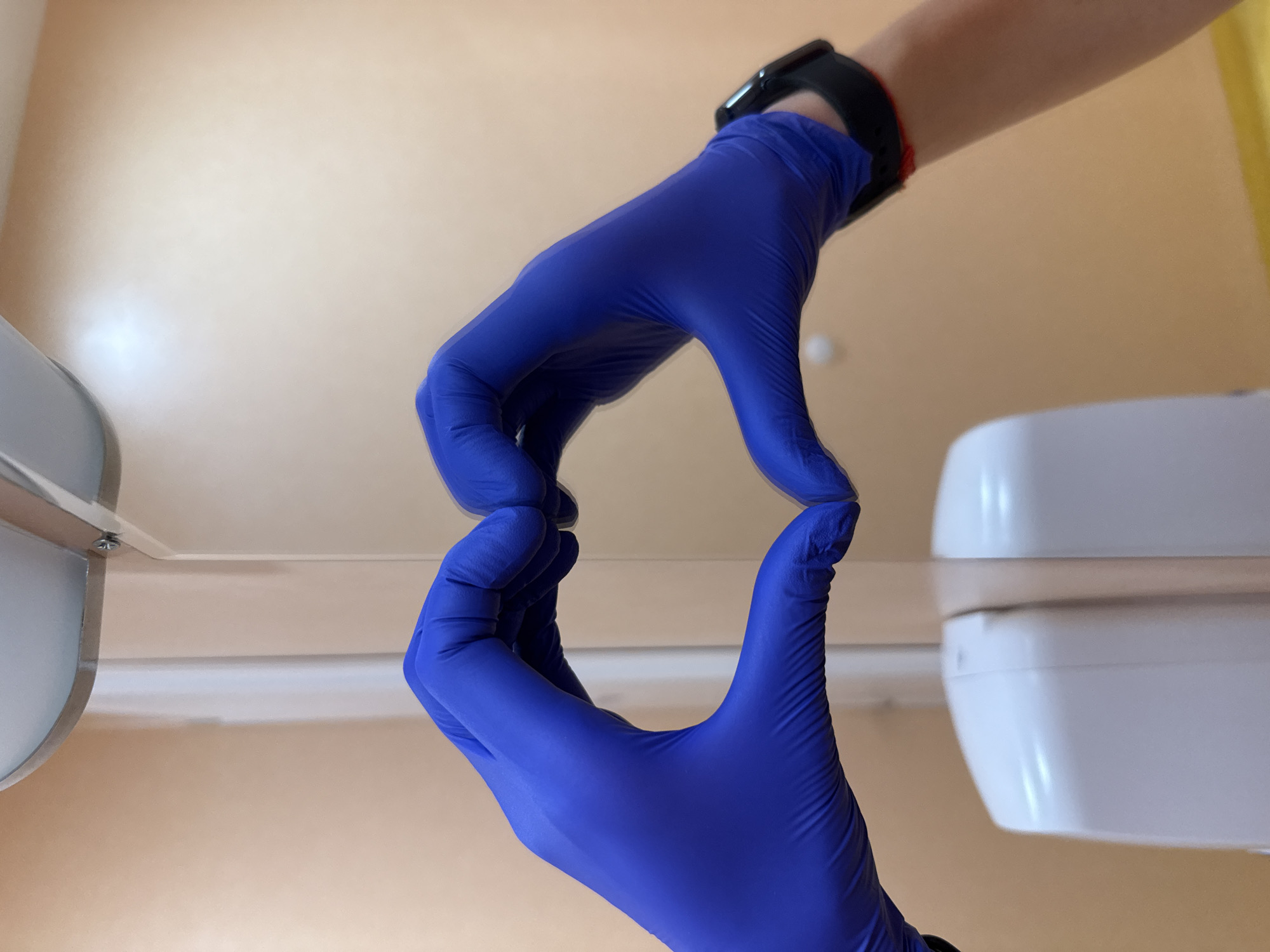 A low-angle, close-up shot features two hands, both covered in bright blue protective gloves, holding a transparent, rectangular sheet of material, likely plastic or glass. The upper hand, connected to a wrist adorned with a black smartwatch, grips the top edge of the sheet, while the lower hand supports it from underneath. The scene's background is a light beige-colored wall or surface. Elements of medical or laboratory equipment are visible on either side: a curved metallic object on the left and a white, angular fixture on the right. The image suggests an indoor clinical, dental, or laboratory setting, possibly in Pleven, Bulgaria, focusing on a precise action, such as examining a sample or preparing for a procedure. No specific time of day, weather, or emotions are discernible, and no text is visible.