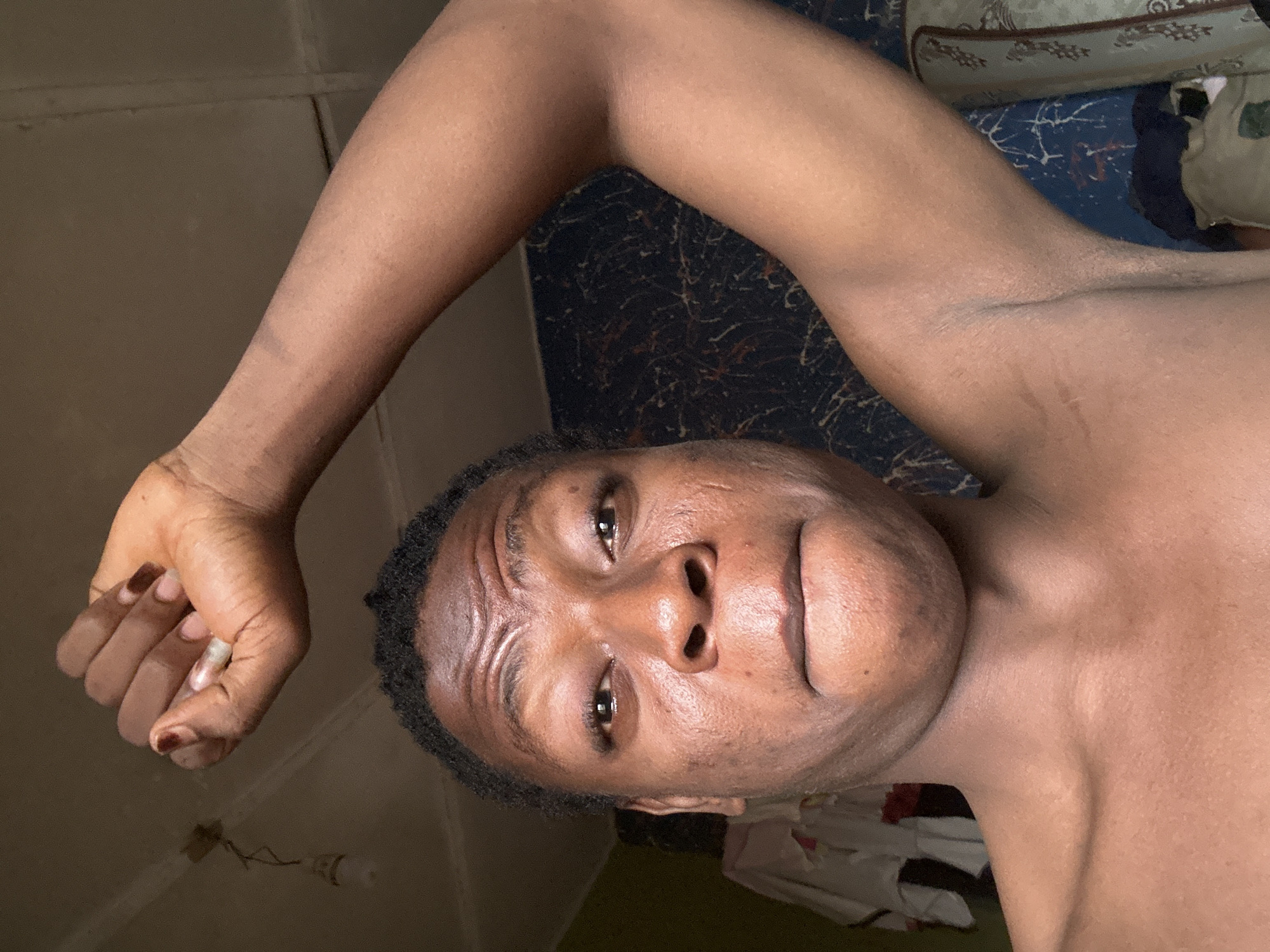 A man with dark skin, short black hair, and brown eyes is lying on his back, facing upwards, with his left arm raised above his head. He appears to be inside a room in a building, possibly a bedroom. The room has a dark background, and there is a bed with patterned bedding behind him. A power cord is visible near his hand. The location in the city is Unknown, Unknown.