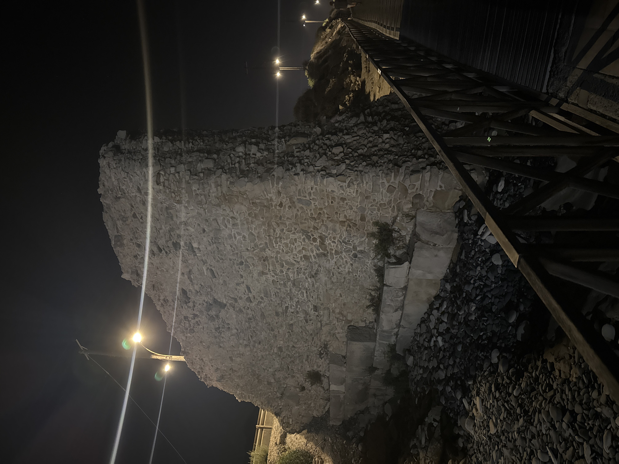 The image depicts a nighttime scene featuring a section of an ancient stone wall, likely part of a historical ruin, situated in the Agios Tychona Community, Cyprus. The wall is constructed from irregular stones of varying sizes, with larger, more squared stones forming the base. The wall is illuminated by streetlights, which cast strong light and shadow, highlighting its textured surface and ruggedness.
To the right of the wall, a wooden boardwalk or walkway with a dark wooden railing extends into the background. The boardwalk appears to be elevated and is positioned over a rocky or pebbled area. In the distance, along a slightly elevated embankment, several streetlights are visible, illuminating the path and surrounding vegetation, which appears dry and sparse. One streetlight is directly behind the large stone wall, its light partially obscured by the structure.
The sky is dark, indicating nighttime. There are no prominent figures visible in the immediate foreground or on the wall itself. However, a silhouette of a person is visible on the boardwalk in the far distance, walking away from the camera. The overall atmosphere is one of a historical site illuminated at night, with a modern walkway providing access or a viewing path. The lighting creates a somewhat dramatic and atmospheric effect.
