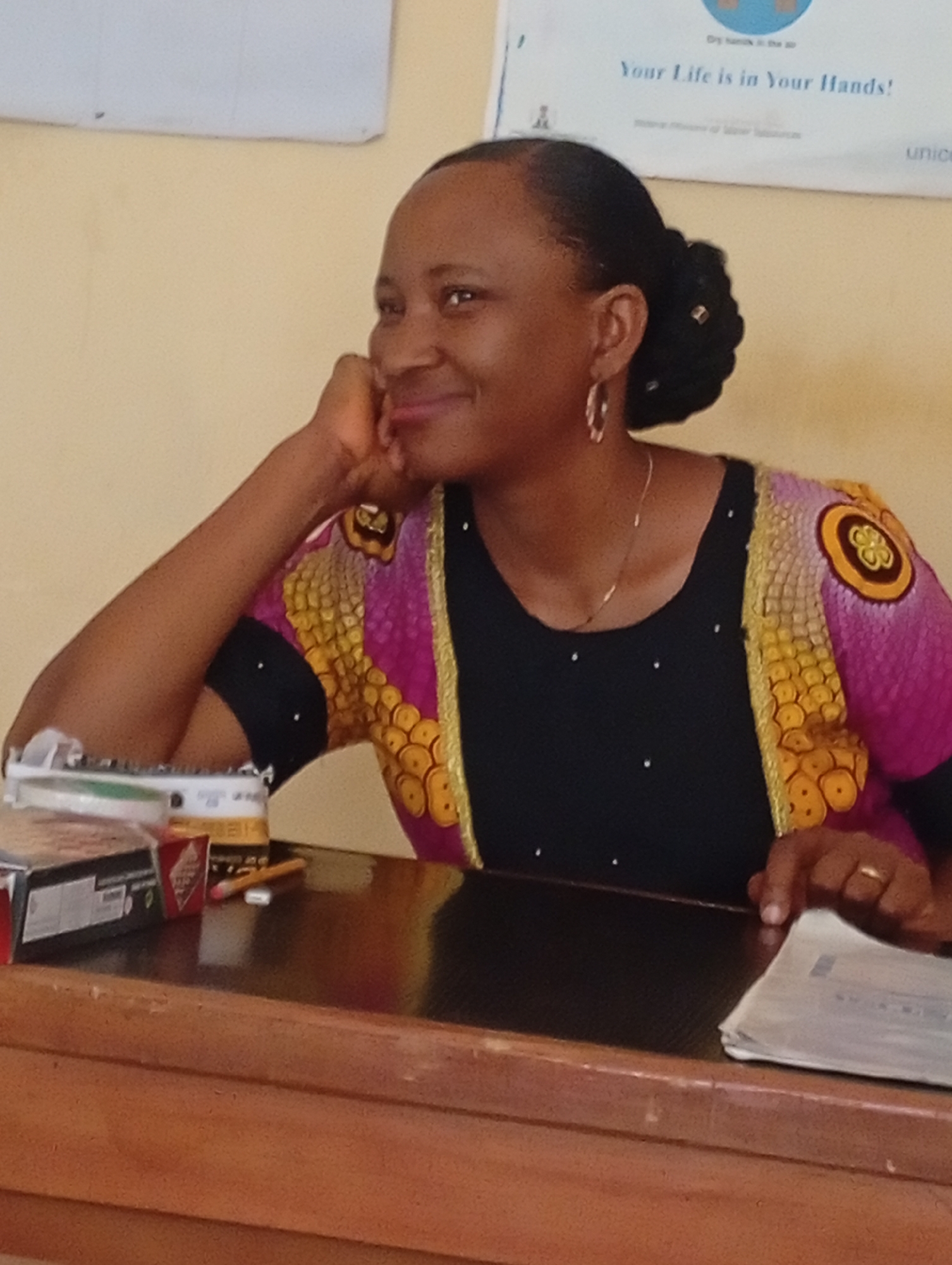 The image shows a woman sitting at a desk. She is wearing a black top with decorative sleeves that are pink, yellow, and black. She has her hair up in a bun and is wearing earrings and a necklace. She is resting her head on her hand and smiling. On the desk, there are boxes, a pencil, and papers. Behind her, there is a sign that reads "Your Life is in Your Hands!" and mentions UNICEF. The location is Unknown, Unknown.