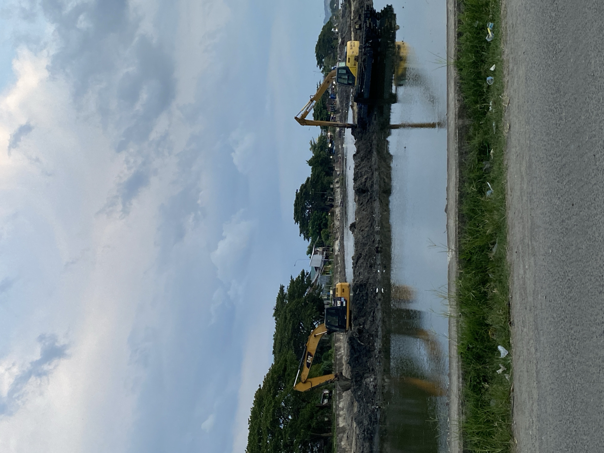 The image depicts an outdoor construction scene by the water, likely in Lhokseumawe, Indonesia. Two yellow excavators are visible in the water, engaged in earthmoving activities. One excavator, a CAT 320D, is positioned on a mound of mud, its arm extended. The second excavator, also yellow and partially submerged, is situated further to the right, with its tracks visible. The water reflects the sky and the machines.
In the background, a line of lush green trees separates the water from a settlement of buildings with visible roofs. A clear sky with scattered clouds dominates the upper portion of the image. In the foreground, a paved road runs alongside a strip of grass, with some litter present on the grass and along the road. The overall impression is one of ongoing development or land reclamation in a coastal area.