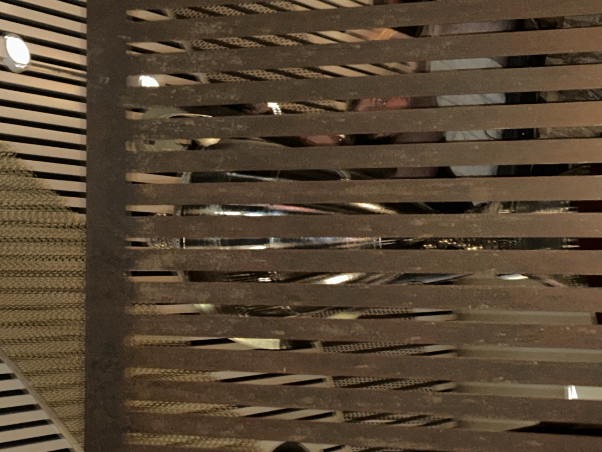 The image depicts a close-up view of a textured metal grate in the foreground, with vertical bars that appear weathered and possibly rusty. Behind the grate, glimpses of an indoor environment are visible, including reflective metallic objects, possibly part of a decorative element or art installation, and segments of a patterned fabric or screen. The ceiling above the grate is adorned with parallel black and white stripes. A spotlight is visible on the ceiling. The scene appears to be taken from a low vantage point, looking upwards through the grate. It is impossible to determine the specific location in Tel Aviv, Israel, or any actions or interactions occurring in the scene due to the obscured view.