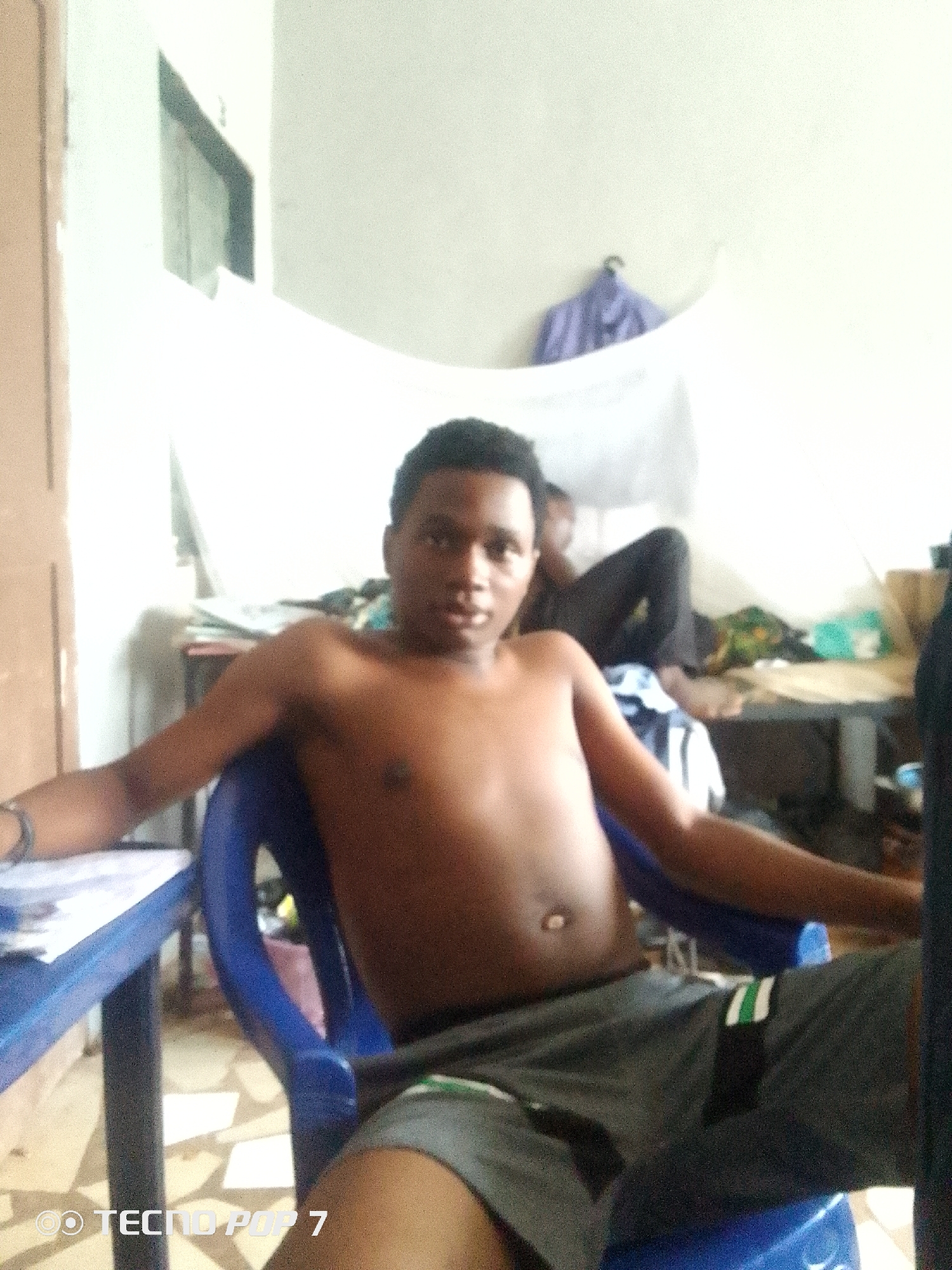 The media file shows an indoor scene with two young men. The primary subject, in the foreground, is a shirtless Black male sitting in a blue plastic chair, facing the camera. He has short dark hair and is wearing grey athletic shorts with green and white stripes. His arms are resting on the chair's armrests, and he appears relaxed. To the right of the primary subject, another Black male is partially visible, lying down in the background, also shirtless and wearing dark pants.
The setting appears to be a sparsely furnished room, possibly a dormitory or a shared living space. In the background, there is a bed or mattress, a table with some items on it, and a white sheet or curtain hanging from the ceiling. A purple shirt is hanging on a hook on the wall. The lighting suggests it is daytime, with natural light coming from an unseen window. The overall atmosphere is casual and informal.
The primary subject is looking directly at the camera with a neutral expression. No specific activity or event is clearly depicted, other than the subjects resting in the room.
The bottom left corner of the image contains visible text: "© TECNO POP 7".
The location context provided is Ipetumodu, Nigeria.