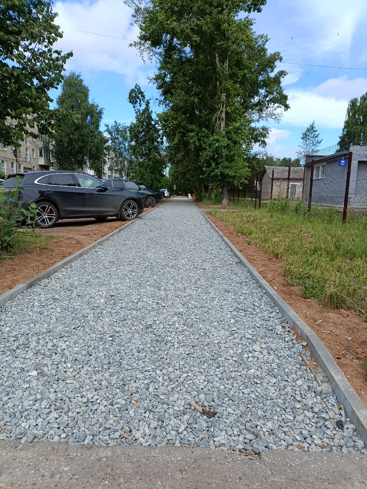 The image depicts a newly constructed gravel pathway in the city of Chaikovsky, Russia, adjacent to a residential building and a fenced area. The pathway is composed of light gray crushed stone, bordered by concrete curbs. To the left of the pathway, several dark-colored cars are parked along the side of the residential building, which has a brick facade and balconies. Mature trees with lush green foliage line the area between the pathway and the buildings, providing shade. To the right of the pathway, a strip of dirt is followed by a grassy area enclosed by a metal mesh fence. Beyond the fence, there are low-rise buildings, one with a gray facade and windows, and another with a weathered concrete or stone exterior. The sky is blue with scattered white clouds. No people are visible in the scene. The overall impression is one of urban development and landscaping.