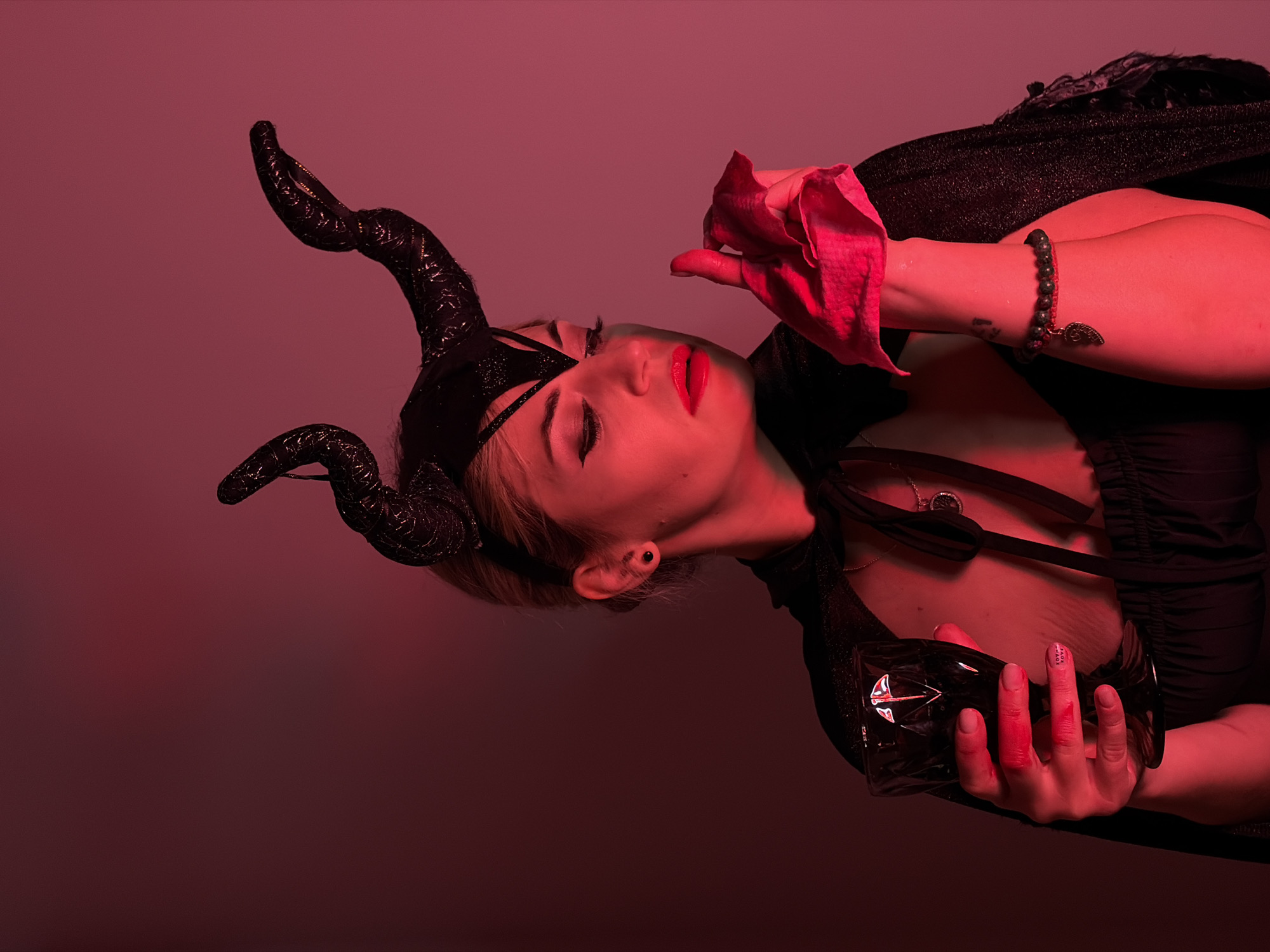 A portrait shot in front of a dark pink background depicts a woman in a costume. She is wearing a black horned headpiece, a black mask over her left eye, and a dark outfit. Her right hand holds a red cloth, while her left hand grips a black glass. She has dark makeup around her right eye and bright red lipstick. A bracelet adorns her right wrist, and her fingernails appear smeared with a red substance. The photo is rotated slightly counter-clockwise. No specific details indicate a location in Budva, Montenegro.
