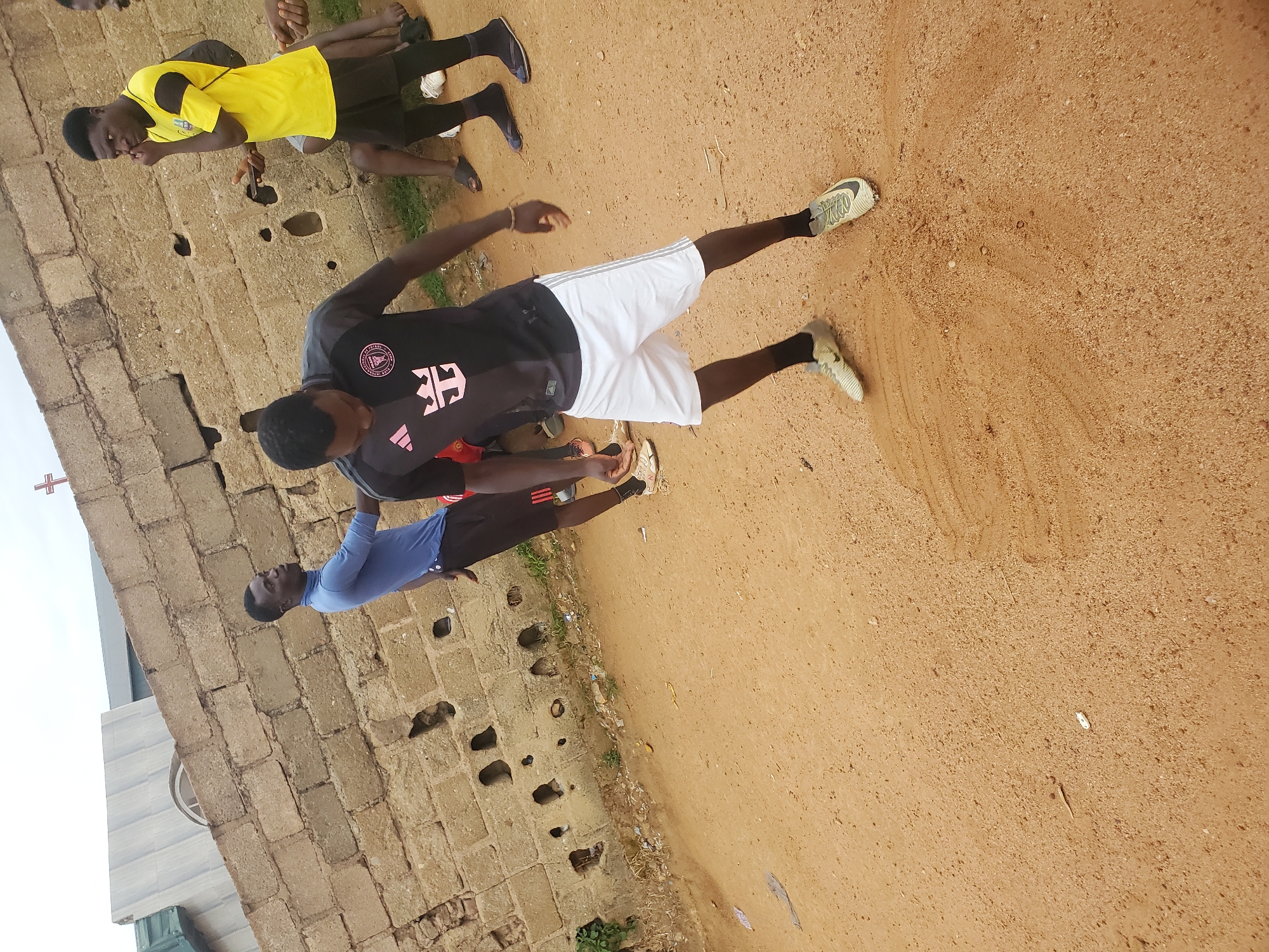 The image shows a group of young men in what appears to be a public area in Jos, Nigeria. The ground is a reddish-brown, sandy dirt. A rough, unfinished brick wall borders one side of the area. The group includes individuals wearing casual athletic attire; one is in a black soccer jersey with white shorts, another in a yellow jersey and black shorts, and another in a blue athletic shirt. They appear to be stretching or warming up. There is a church or large building with a cross visible in the background.