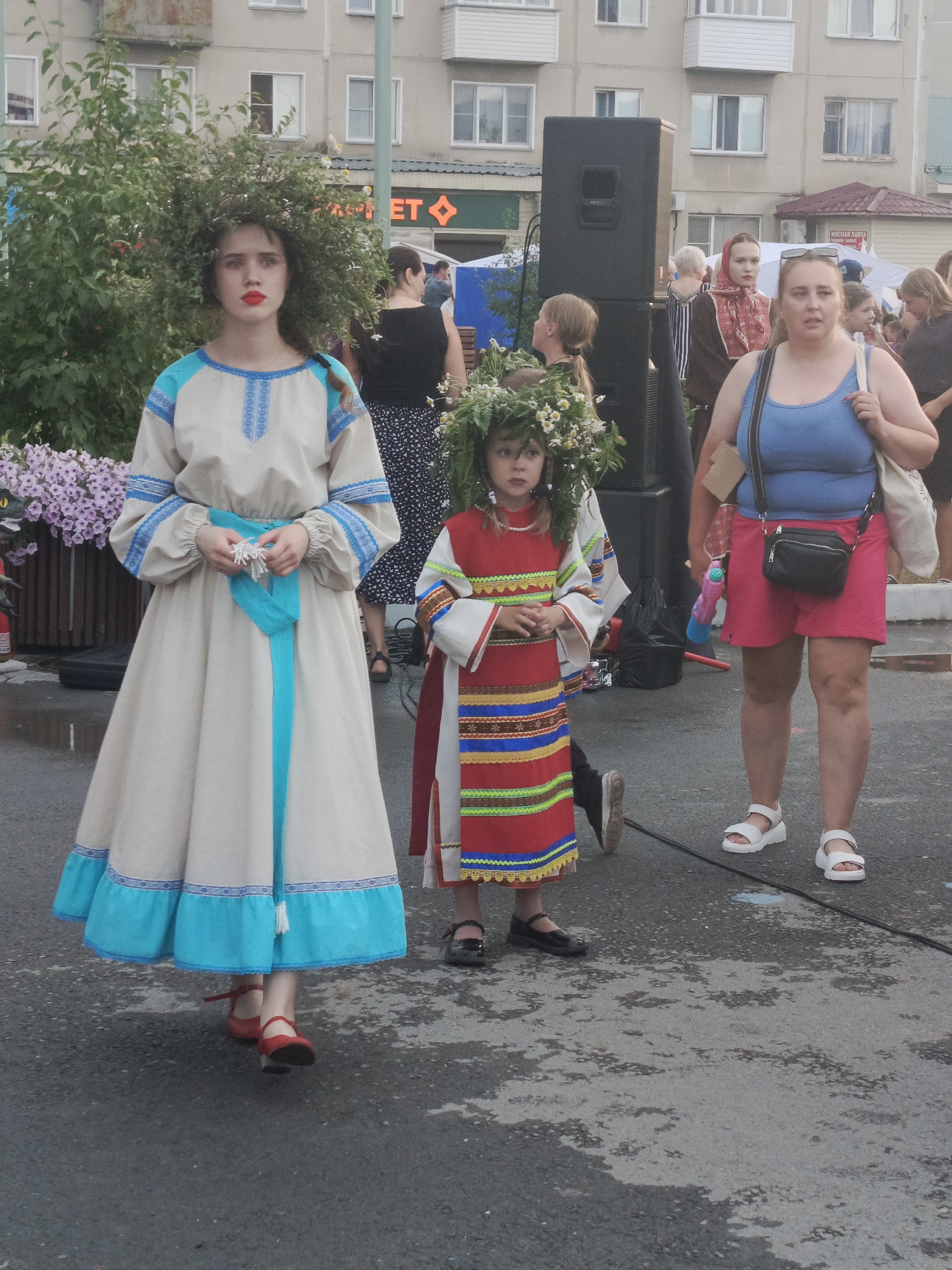 The image depicts a daytime outdoor gathering in Kryvdaivka, Russia, likely a festival or cultural event, given the traditional attire of two young girls. In the foreground, a young girl with a large, leafy floral wreath on her head and a colorful, patterned dress stands facing the viewer. To her left, an older girl, also adorned with a voluminous green floral wreath, wears a cream-colored, long-sleeved dress with blue embroidery and trim, and red shoes. Her expression is serious.
Behind them, a woman in a blue tank top and pink shorts, wearing white sandals, stands looking off to the side. A black crossbody bag is slung over her shoulder. Other people are visible in the background, including a woman in a red headscarf and a person in a black top and patterned skirt. A large black speaker is positioned to the left of the woman in the pink shorts. The setting appears to be a paved area in front of a multi-story building, with some greenery and flowers present. The lighting suggests it is daytime, and the weather appears overcast, though not actively raining. The mood seems to be one of community celebration. There is visible text on a building in the background, part of which appears to say "ET".