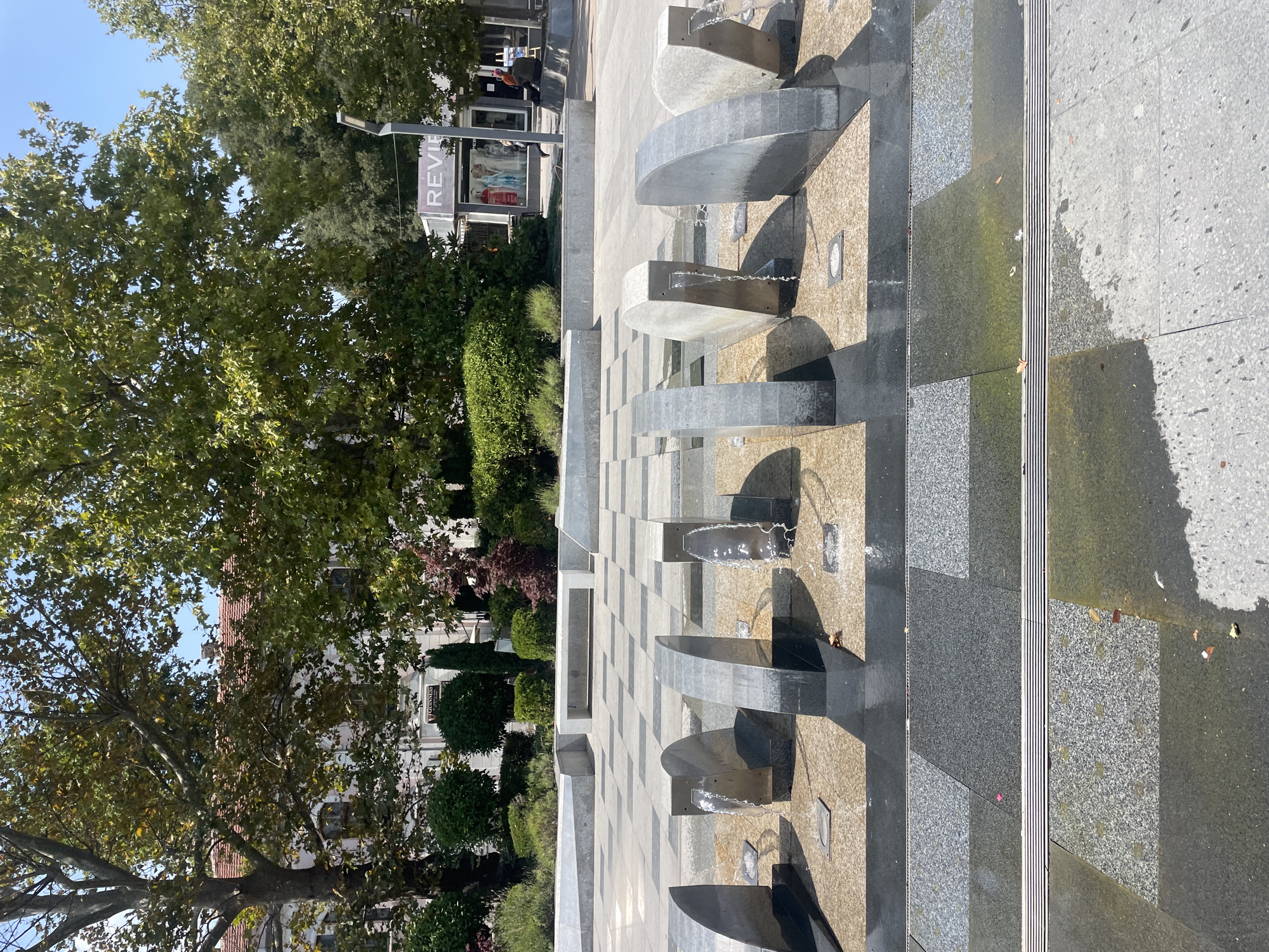 The image depicts a contemporary public space with a water fountain feature in the foreground. The fountain consists of several modern, curved stone elements from which water streams down. The ground around the fountain is paved with dark and light granite tiles in a checkered pattern, with some areas wet from the water.
In the mid-ground, there are low stone benches and planters with manicured shrubs and trees. Behind these, a building with a red-tiled roof and white facade is visible, partially obscured by large trees with dense green foliage. Further in the background, a shop with a prominent "REV...A" sign is situated. A street lamp is also present. One person is visible walking in front of the shop, and two people are seated on a bench in the distance.
The scene is set outdoors on a sunny day, with a clear blue sky. The overall impression is of an urban public square designed for relaxation and enjoyment.
This location is the "Velikost" square (also known as "The Sea Garden Boulevard"), located in the city of Burgas, Bulgaria.