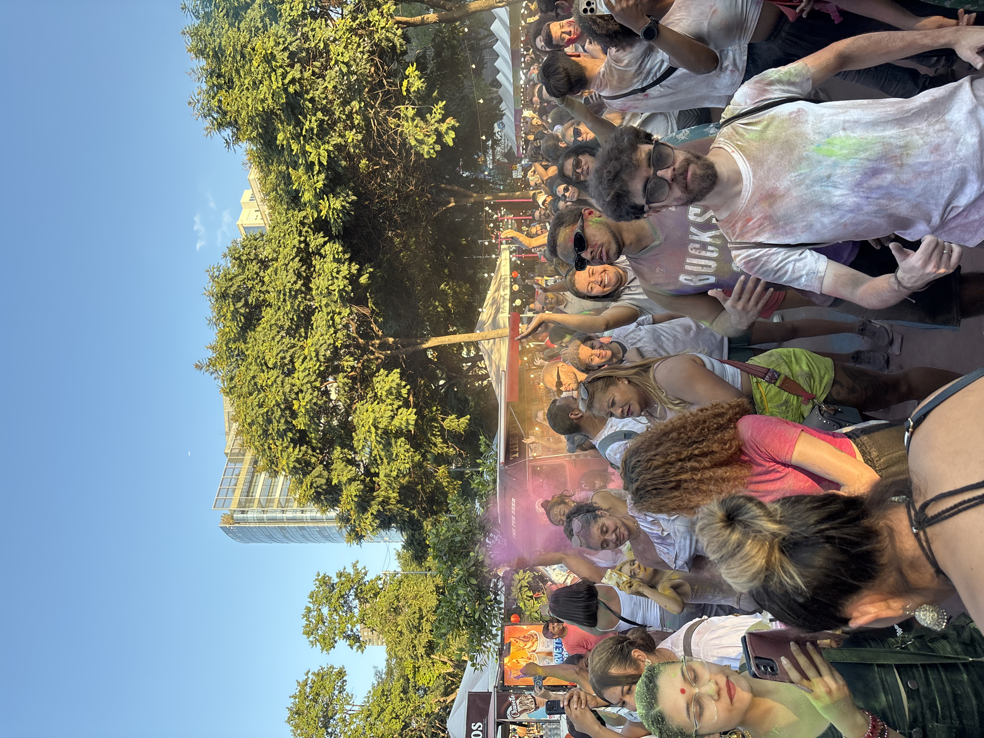 The image shows a large crowd of people gathered outdoors, likely at a festival or event in the city of São Paulo, Brazil. Many are covered in colored powder, suggesting a Holi festival celebration. The crowd is dense and appears to be enjoying the event, with some people smiling and raising their hands in the air. Some are taking photos or videos. The event is located in a park with tall trees and a modern high-rise building visible in the background. There are vendor booths or stands along the periphery. The sky is clear and blue, indicating a sunny day.