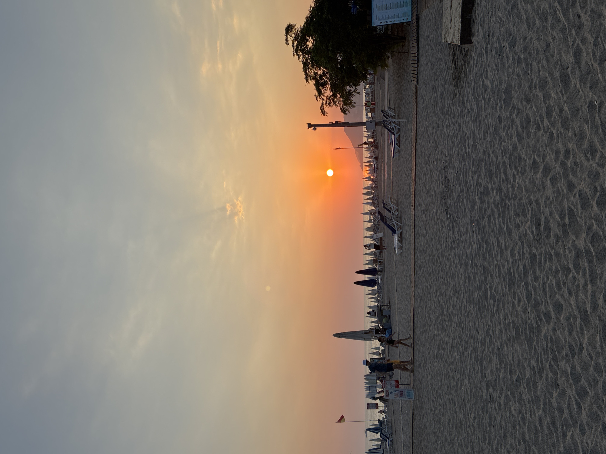 The image depicts a sunset at Cleopatra Beach in Alanya, Turkey. The sun, a bright orange orb, is low on the horizon, casting a warm glow across the sky and illuminating the clouds with hues of orange and yellow. The beach is covered in sand, with rows of empty lounge chairs and closed umbrellas spread out in front of the sea. A few people are visible on the beach, including a man and a child walking in the foreground, and other figures scattered further down the beach. A red and yellow flag is planted on the left side of the beach. The sea is calm and reflects the colors of the sunset. The overall scene conveys a sense of tranquility and the end of a day at the beach.