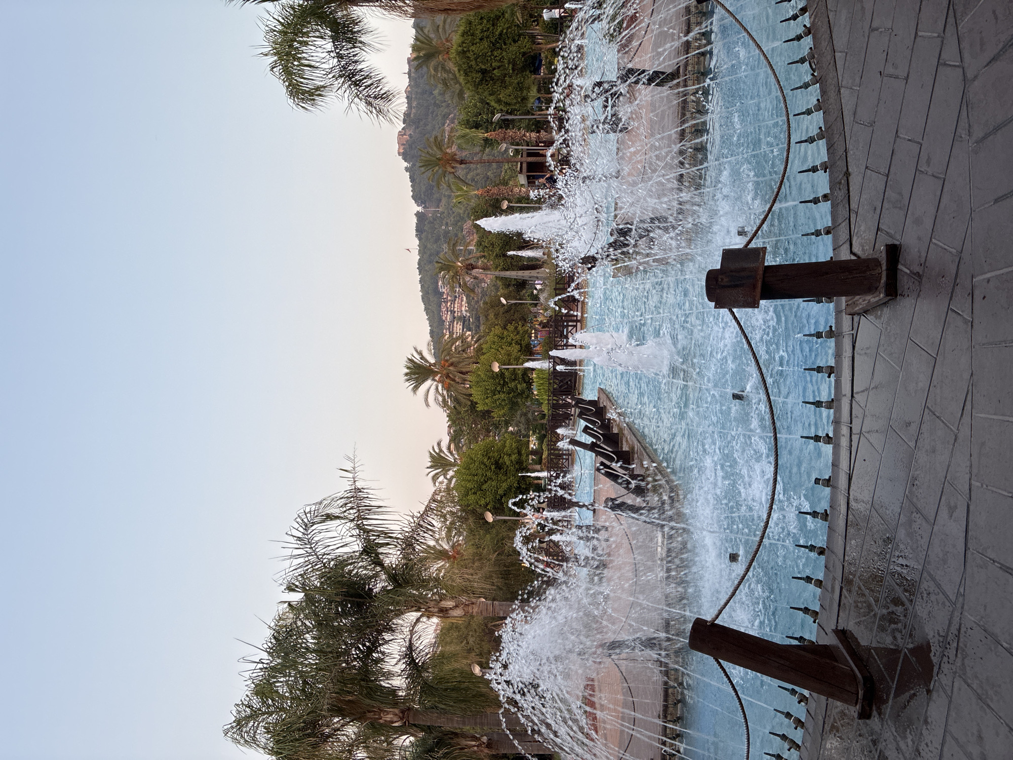 This image depicts a public fountain area in Alanya, Turkey. In the foreground, a wide fountain sprays water upwards and outwards, creating a misty effect. The fountain is situated in a shallow pool with bright blue water. The surrounding area is paved with dark grey interlocking tiles, some of which appear wet. A rope barrier, supported by wooden posts, separates the paved area from the fountain. In the middle ground, a series of smaller fountains are visible, as well as a wooden boardwalk with railing. Palm trees and other lush green vegetation are abundant throughout the scene, some of which are in the middle ground and background. In the background, a hill is visible with buildings and a castle-like structure at the summit. The sky above is a clear, pale blue, suggesting it is either early morning or late afternoon. Several individuals are casually walking or standing in the background, seemingly enjoying the park. The overall impression is that of a well-maintained public space designed for leisure and recreation.
