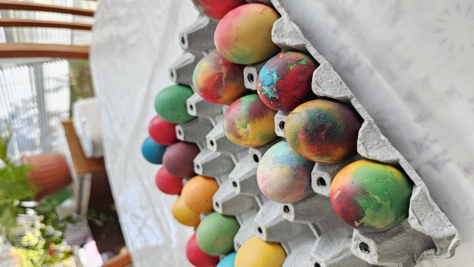 A close-up, high-angle shot depicts Easter eggs in a gray, cardboard egg carton. The eggs are dyed with multicolored patterns. The carton is positioned against a white cloth. To the left, the scene blurs, revealing a partial view of a room with a plant in a brown pot, and a window. The location is not identifiable as Varna, Bulgaria.