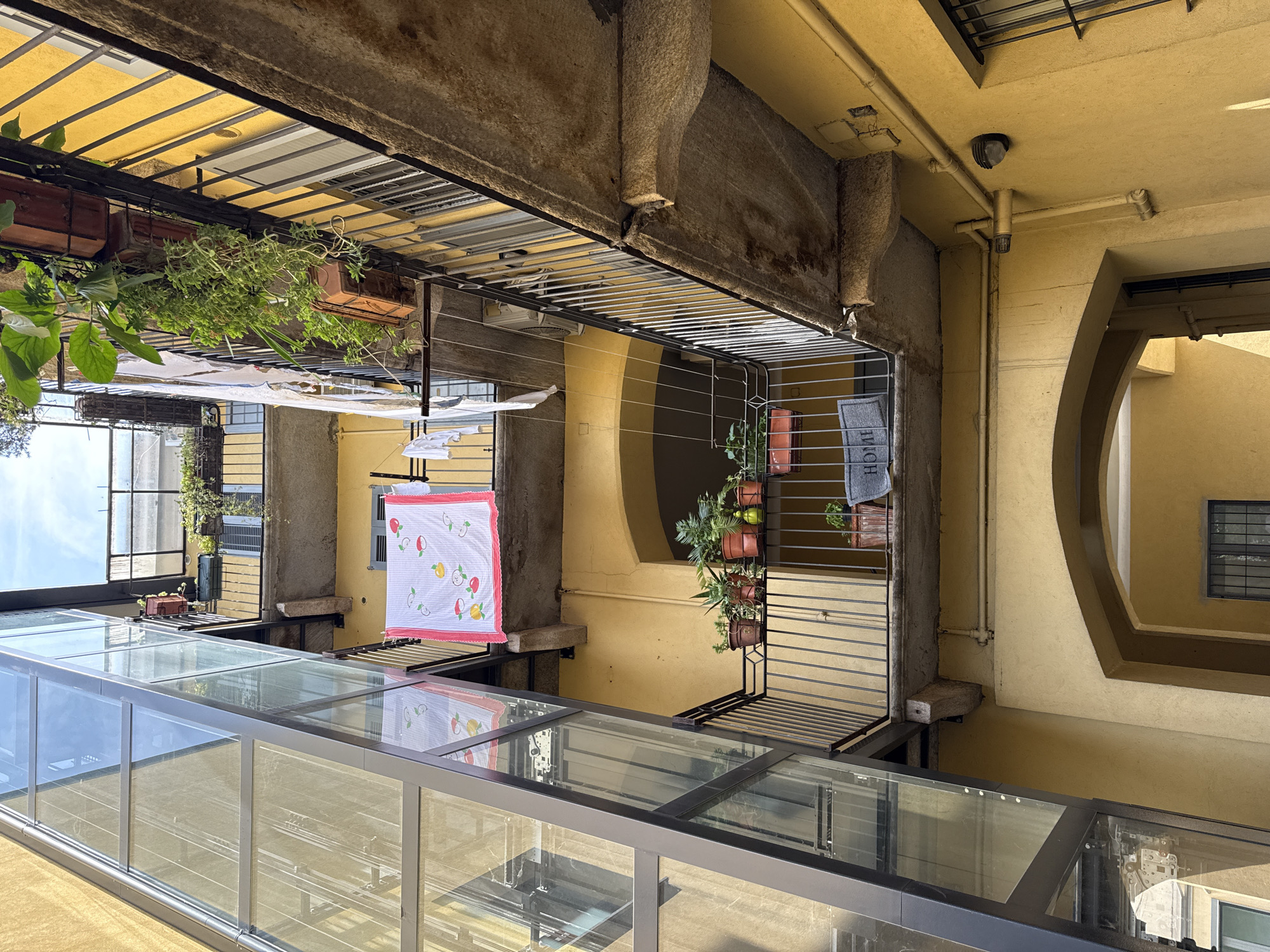 This is a low-angle, full shot of the inner courtyard of a building in Milan, Italy, featuring a modern glass elevator shaft on the left and multiple levels of balconies with railings and potted plants. The building's facade is painted a bright yellow. Laundry is hung on clotheslines on some of the balconies. On the ground floor, an arched entryway is visible. The lighting suggests it is daytime, possibly late morning or early afternoon, with sunlight casting shadows.