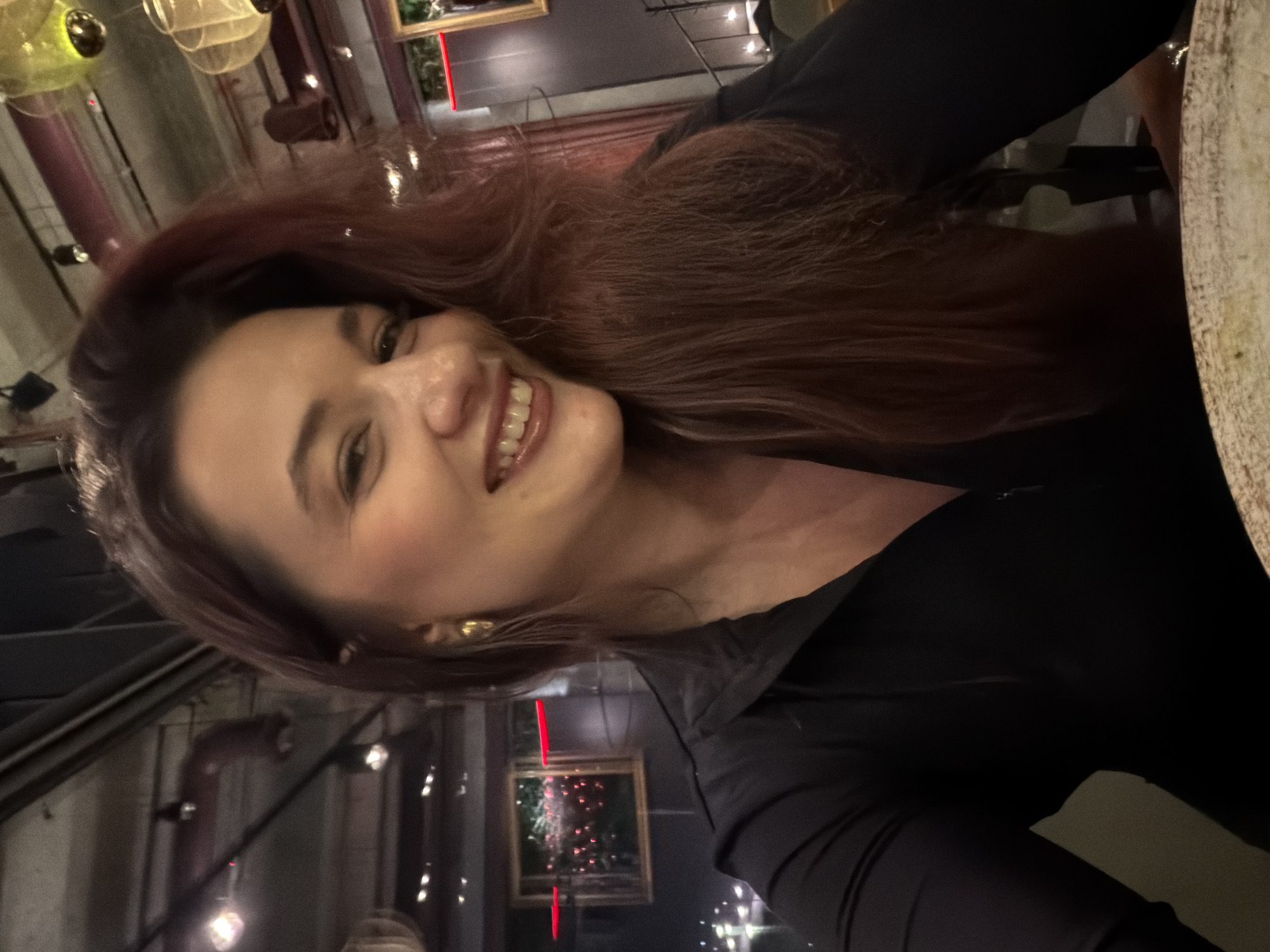 A female individual is depicted from the chest up, positioned centrally in the frame, looking directly at the camera with a broad smile. She has long, dark reddish-brown hair, styled with volume. Her attire consists of a black, long-sleeved top with a zippered neckline. She is wearing gold-colored, elongated drop earrings. A light-colored, textured plate is partially visible in the immediate foreground, suggesting she is seated at a table.
The background reveals a dimly lit interior with an industrial-chic aesthetic. The ceiling features exposed dark structures, possibly pipes or ventilation, along with track lighting and various suspended pendant lights, some appearing amber or green. Behind the subject, a large window or glass panel reflects blurred exterior city lights, including distinct red and white light points, and a prominent horizontal red light strip. Framed elements containing greenery or abstract patterns are visible on the wall behind the glass. Red curtains or drapes are partially visible on the far right. The overall environment suggests a modern restaurant or bar.
The location is identified as Belgrade, Serbia, specifically consistent with the interior design, lighting scheme, and reflected urban landscape of Buddha-Bar Bistro Belgrade.