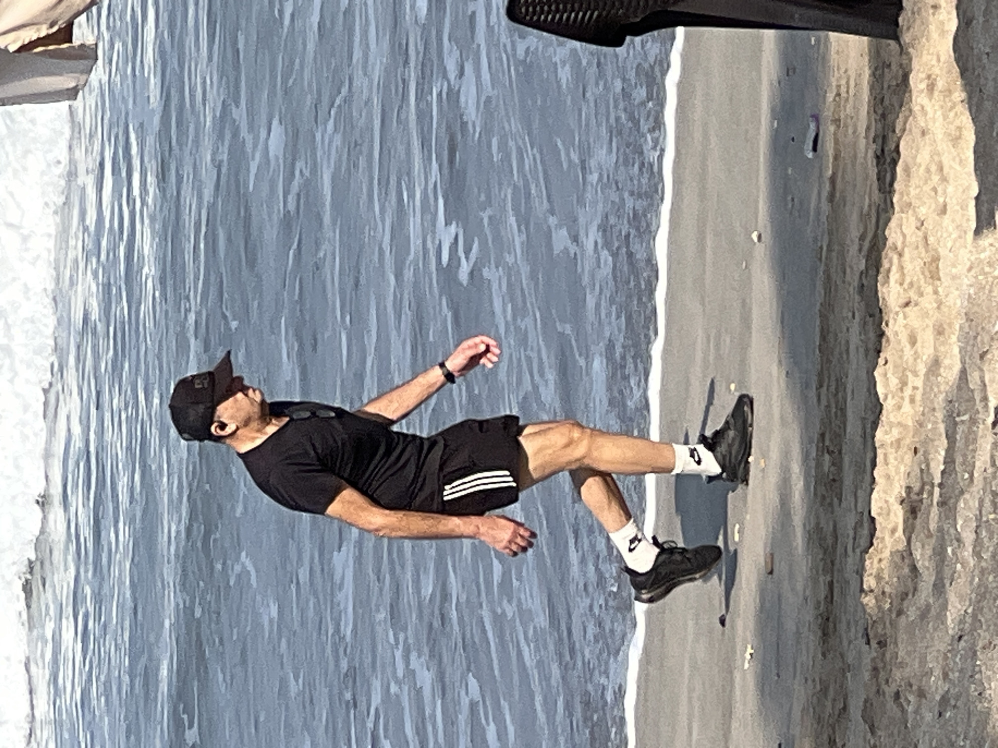 A man wearing a black t-shirt, black shorts with white stripes, white socks, and black athletic shoes walks along the shoreline on a sandy beach. He also wears a black baseball cap and appears to have sunglasses clipped to his t-shirt. The ocean is to his left with gentle waves lapping at the shore. To his right, a dark woven chair is partially visible. The environment suggests a beach setting in Kuta, Indonesia.