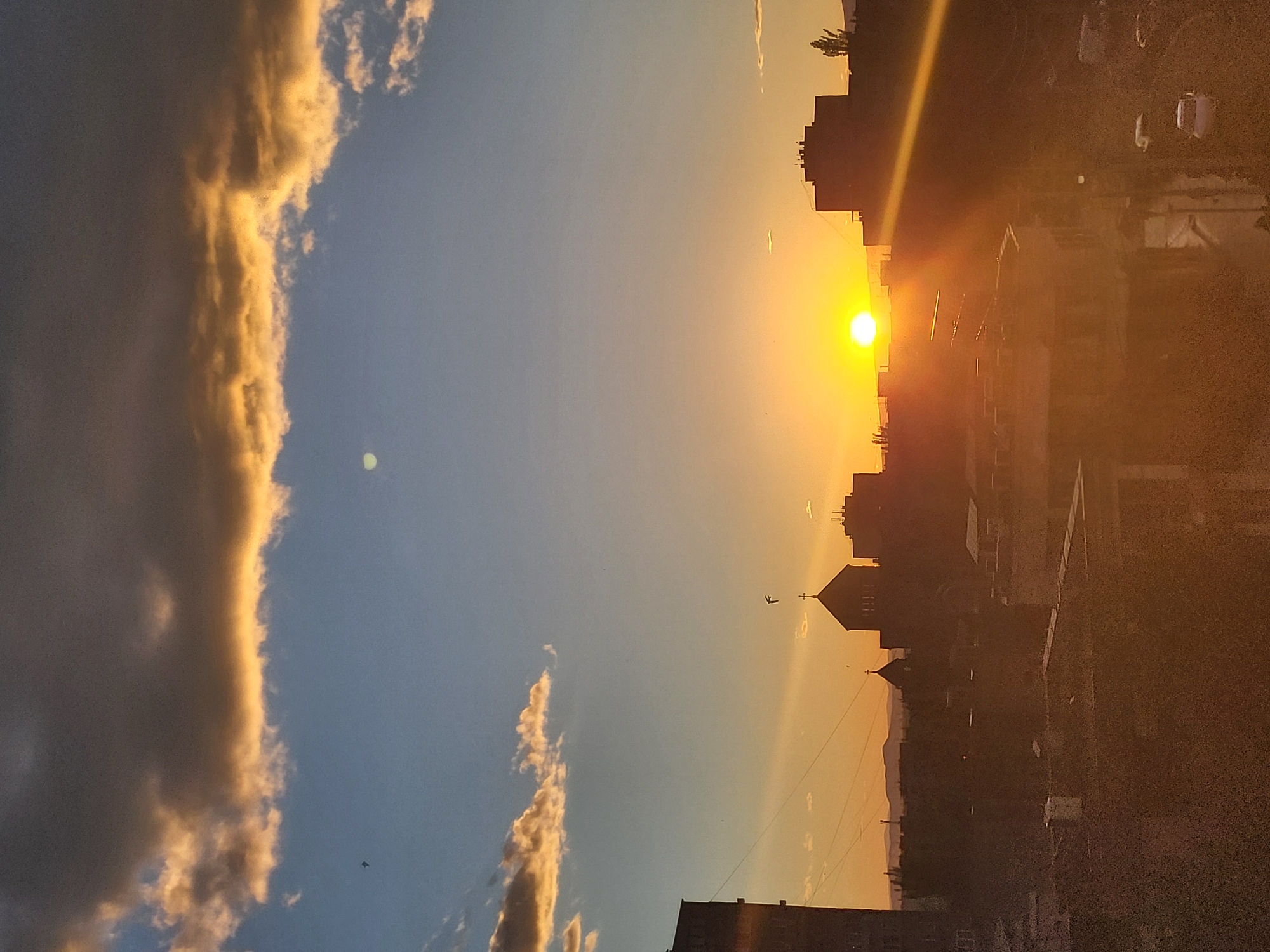 The image depicts a sunset in Yerevan, Armenia, with the sun positioned at the horizon, partly obscured by building silhouettes. The sky transitions from a bright yellow near the sun to a pale blue. Buildings, some with distinct features like a church spire and multiple stories, are silhouetted against the bright sky. Clouds, illuminated by the setting sun, stretch vertically along the left side of the image. Power lines cross the sky. A small aircraft flies in the sky near the spire.