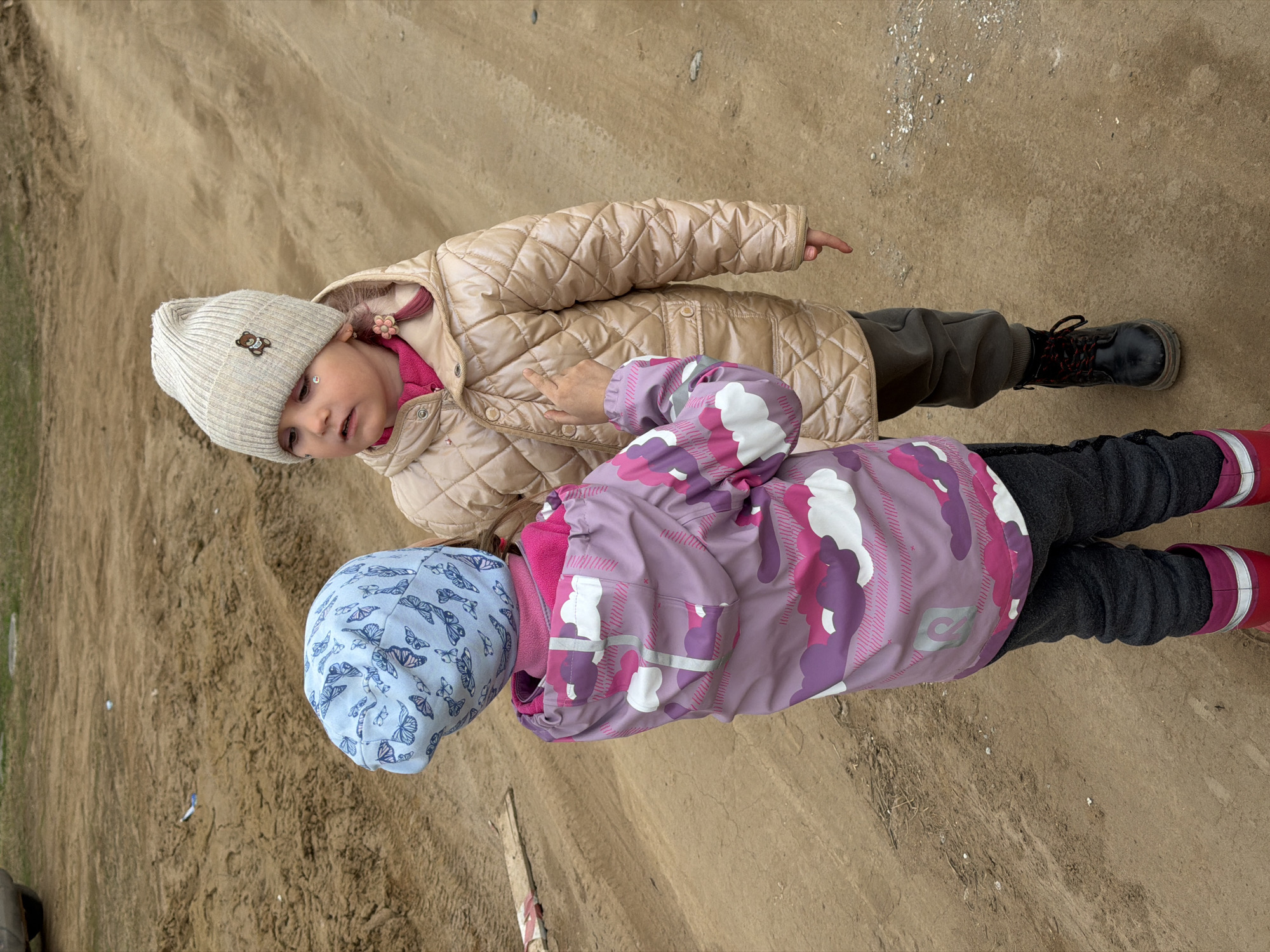 Two young children stand on a sandy surface. The child in the foreground wears a blue hat with a butterfly pattern and a purple, pink, and white patterned jacket with grey pants and red boots. The second child wears a light beige quilted jacket with a matching hat adorned with a small bear emblem, dark-colored pants, and black boots. Both are facing the camera, with their fingers pointing towards something out of frame. The sandy ground appears to be a mixture of sand and dirt, common in outdoor recreational areas. The location is Chaikovsky, Russia.
