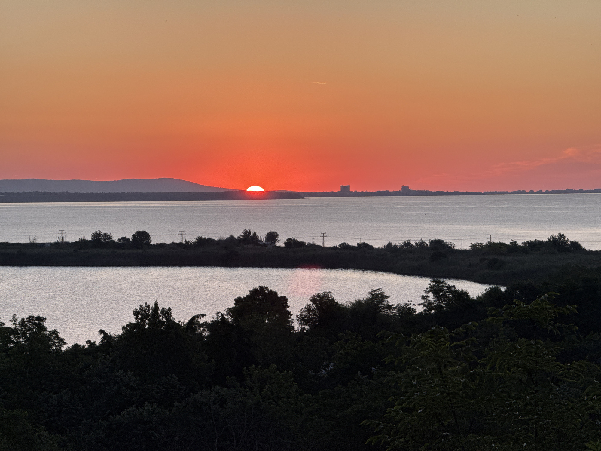 The image captures a sunset over the Burgas Bay in Burgas, Bulgaria. The sun, a vibrant red orb, is partially submerged below the horizon, casting a warm orange and red glow across the sky. The calm waters of the bay reflect the sunset's colors, creating a serene and picturesque scene. In the distance, a thin strip of land is visible, with a mountainous silhouette on the left and buildings on the right, suggesting the presence of urban development along the coastline. The foreground is dominated by dark silhouettes of trees, their leaves and branches creating a natural frame for the sunset. The overall impression is one of tranquility and natural beauty, a typical evening view from the coastal city of Burgas.