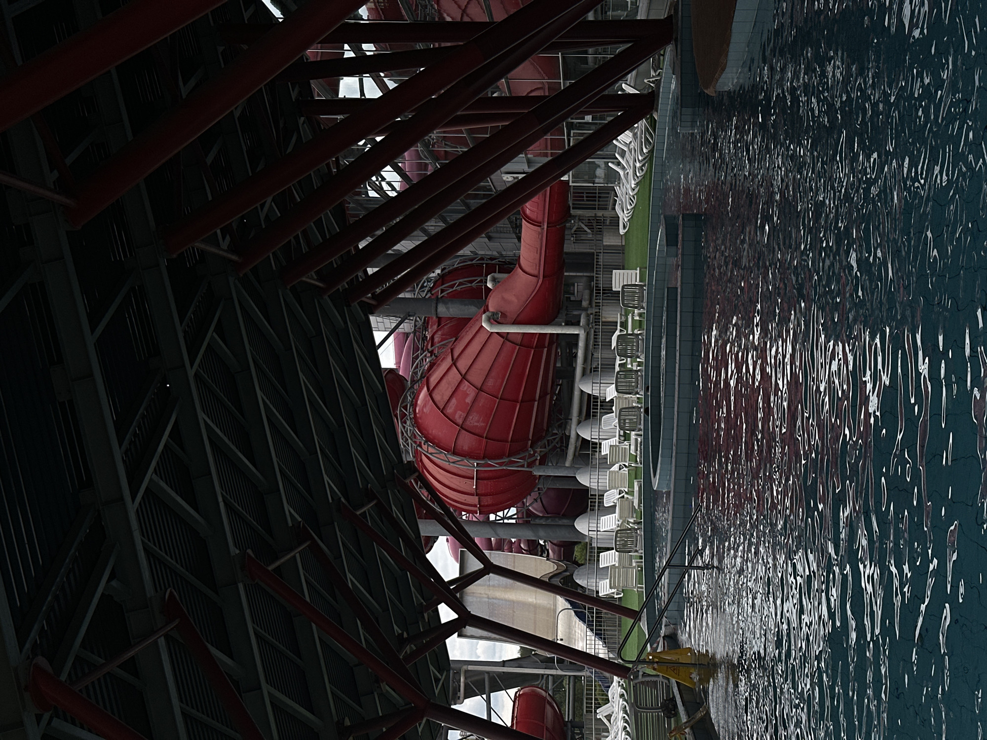 The image depicts an indoor water park in Minsk, Belarus. The primary subjects are the red water slides, a large swimming pool, and rows of white lounge chairs. The scene is an indoor complex with a high ceiling supported by a dark metal structure. The water in the pool is a deep blue-green, with light reflecting off its surface, suggesting artificial lighting. The water slides are prominently featured, with their vibrant red color contrasting with the darker structural elements and the pool water.
The setting appears to be a recreational facility, likely an indoor swimming pool and water park, given the presence of slides and lounge chairs. The overall atmosphere suggests a place for leisure and fun. There are no people visible in the image, nor is there any clear indication of the time of day or specific weather conditions beyond the fact that it is indoors. The location is identified as Minsk, Belarus. There is no visible text in the image. The lighting is somewhat dim in the structural areas, with brighter illumination over the pool and slides. The image is oriented vertically, with the pool and slides extending downwards.