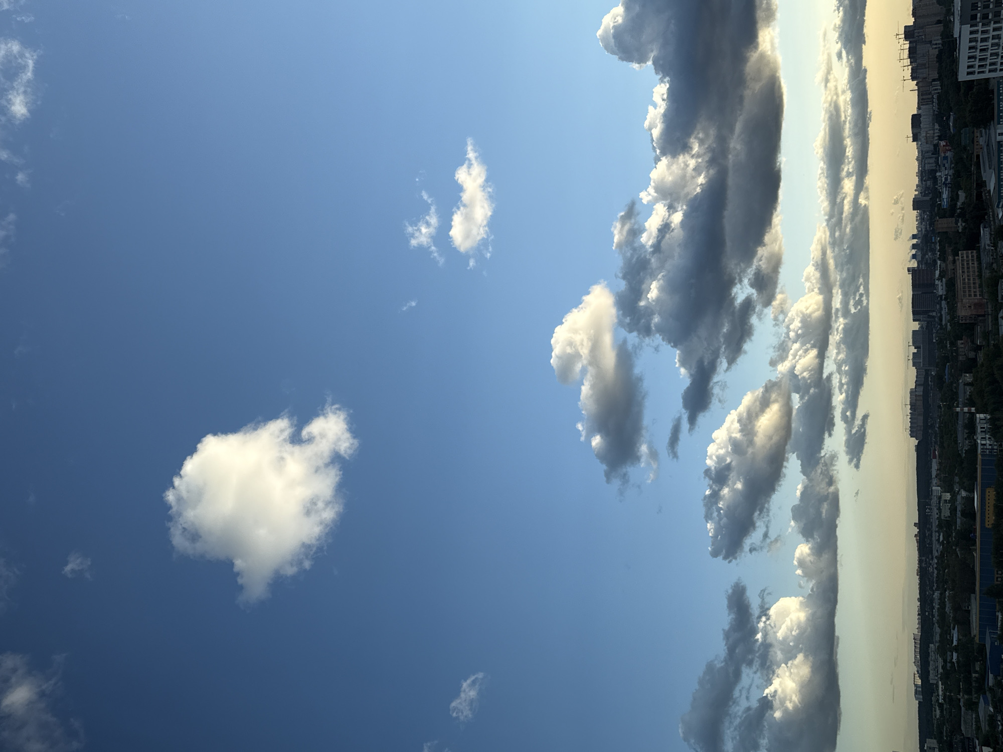 The image shows a wide view of the sky over Lyubertsy, Russia, with a cityscape in the background. The sky is a clear blue with scattered white and grey clouds. Some clouds are illuminated by the sun, appearing bright white, while others are darker and appear to be casting shadows.
In the foreground, the upper portion of the image is dominated by the sky. A large, fluffy white cloud is prominent in the upper left quadrant. Further down, a band of darker, more textured clouds stretches across the middle of the image, some of which are tinged with yellow, suggesting late afternoon or early evening light. A few smaller, wispy clouds are scattered throughout.
The background features the urban landscape of Lyubertsy. Buildings of various sizes and architectural styles are visible, including apartment complexes and industrial structures. Construction cranes can be seen among some of the taller buildings, indicating ongoing development. Green trees and vegetation are interspersed throughout the city, suggesting a mix of residential and natural areas. The horizon line is low, with the city appearing to spread out beneath the vast expanse of the sky. The overall impression is of a city under a dynamic and cloud-filled sky.
