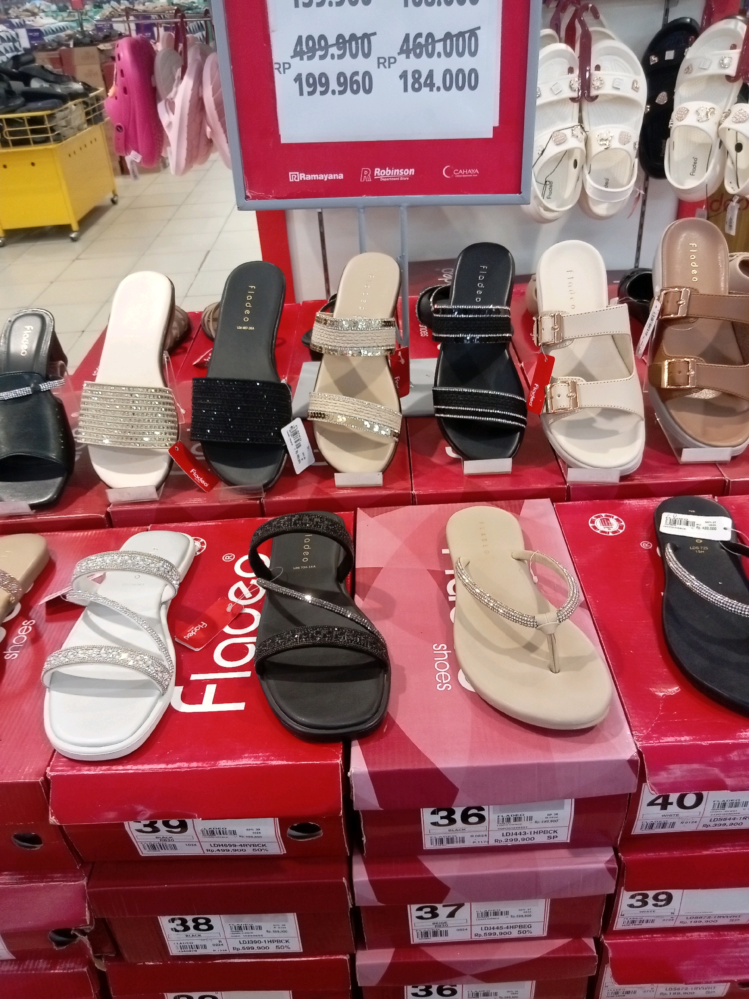 The image shows a retail display of women's footwear, likely in a department store or shoe shop in Batam, Indonesia. Several pairs of sandals and flip-flops are arranged on red display boxes. The footwear features various designs, including embellished straps with rhinestones, metallic accents, and simple flat soles. Some of the shoes are black, white, beige, and brown. Price tags are visible, indicating discounted prices on some items. In the background, more shoes are displayed on racks, and a yellow shopping cart is partially visible. The overall environment suggests a commercial setting with ample merchandise. The context of Batam, Indonesia, implies this is likely a sale or promotional event within a local retail establishment.
