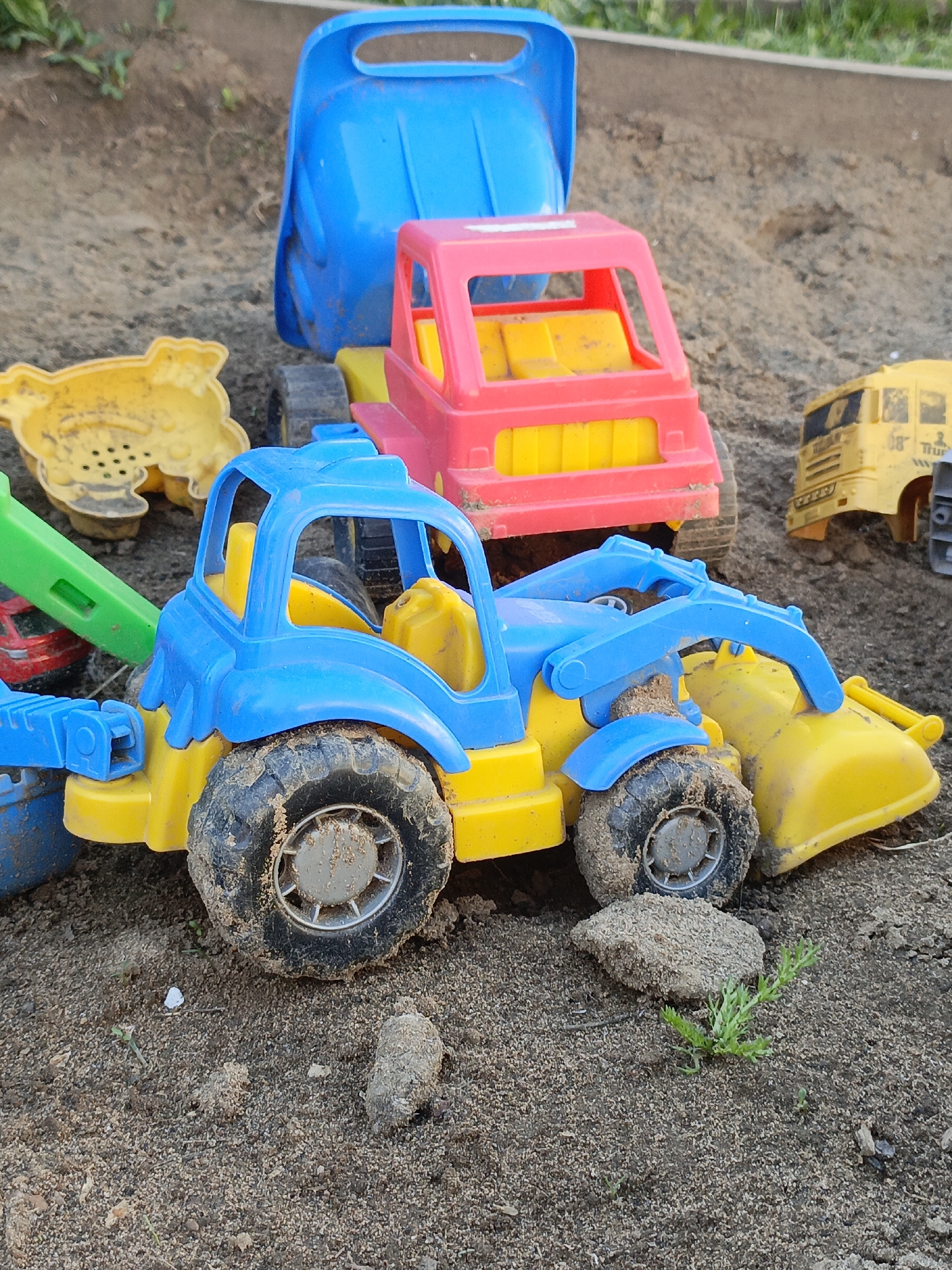 The image shows a collection of colorful plastic toy trucks and other sandbox toys scattered on a sand surface. The primary focus is a blue and yellow front-end loader style toy, covered with sand. Behind it are a pink and yellow truck, and a blue bucket. Other yellow toys are partially visible to the left and right. The toys are set on sand. A few small rocks and some vegetation are present near the base of the front-end loader. There are no people visible in the image. The scene appears to be a children's sandbox or play area. There is no evidence to determine the location or city. The claim that it is in Gabdyukovo, Russia, cannot be verified from the image.