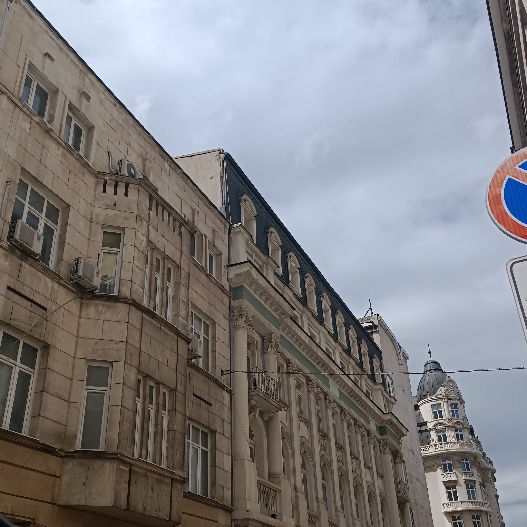 The image captures a street scene in Sofia, Bulgaria, showcasing a collection of multi-story urban buildings under an overcast, cloudy sky during daytime. The perspective is looking upwards at the facades of these structures.
On the left, an older, perhaps stone-clad building is visible, featuring numerous windows and several external air conditioning units mounted on its facade and a bay window section. The building exhibits signs of weathering.
Adjacent to it, towards the center and right, stands a more ornate building, possibly of Neoclassical or similar architectural style. This structure is characterized by decorative columns, balustrades, arched windows, and a prominent dark mansard roof dotted with dormer windows. Its facade shows intricate detailing and a lighter color palette compared to the building on the left. Overhead wires are visible running between these buildings.
Further in the background and to the right, another distinct building rises, crowned by a large, dark dome or cupola with a small spire at its apex. This building also features multiple levels of windows and balconies, displaying elaborate decorative elements consistent with turn-of-the-century European architecture.
In the upper right corner of the frame, a circular street sign is partially visible. It has a red outer border, a white inner ring, and a blue center with white markings, indicating a "no parking" or "no stopping" regulation.
No human activity is depicted in the frame, and no specific text is clearly legible on any of the buildings or the partial sign.