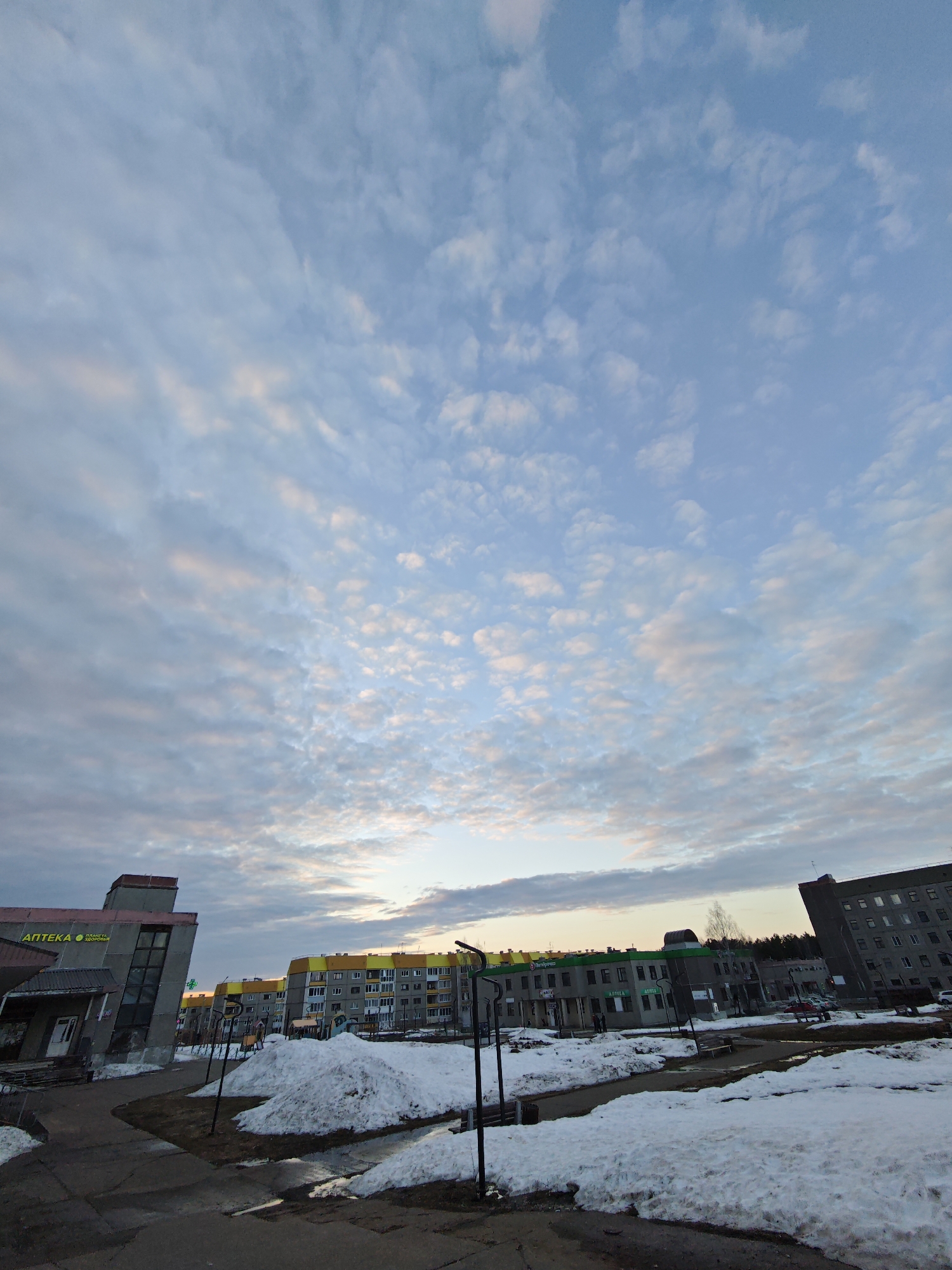 The image depicts a street scene in Markovskii, Russia during the early spring. Patches of melting snow are visible on the ground. Buildings line the street, including a pharmacy labeled "АПТЕКА" in Russian. The sky is filled with scattered clouds. The buildings are mostly concrete, some with green and yellow accents. No people are clearly visible in the scene. The photograph appears to be taken at ground level.