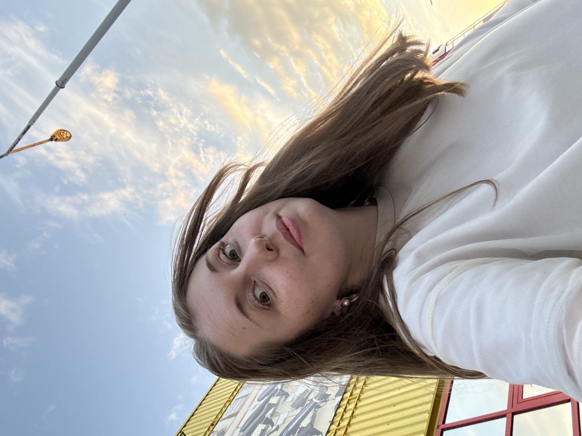 A young adult female is depicted in a close-up, upward-angled selfie, against an outdoor backdrop. She has light brown hair, long and slightly wind-blown, with some strands framing her face. Her complexion is fair with visible freckles, and she has light-colored eyes with a wide-eyed, slightly surprised expression, looking directly into the camera. She wears a single pearl drop earring on her visible left ear. Her lips are a natural pink, slightly parted. She is wearing a loose-fitting, white long-sleeved shirt with a crew neck.
The background on the left features a building with bright yellow corrugated metal siding, below which is a red-framed window or structure reflecting the sky. A large, monochrome graphic or mural, possibly depicting multiple mechanical objects or watercraft, is visible on the yellow siding. To the right, a tall, slender grey pole extends upwards, with an orange, flattened oval object, possibly a light or sensor, attached higher up. The sky is bright blue, transitioning to a warm golden-orange hue on the right, indicative of late afternoon or early evening. Scattered, wispy white clouds are present across the sky. The location is an unspecified outdoor area in the city of Unknown, Unknown.