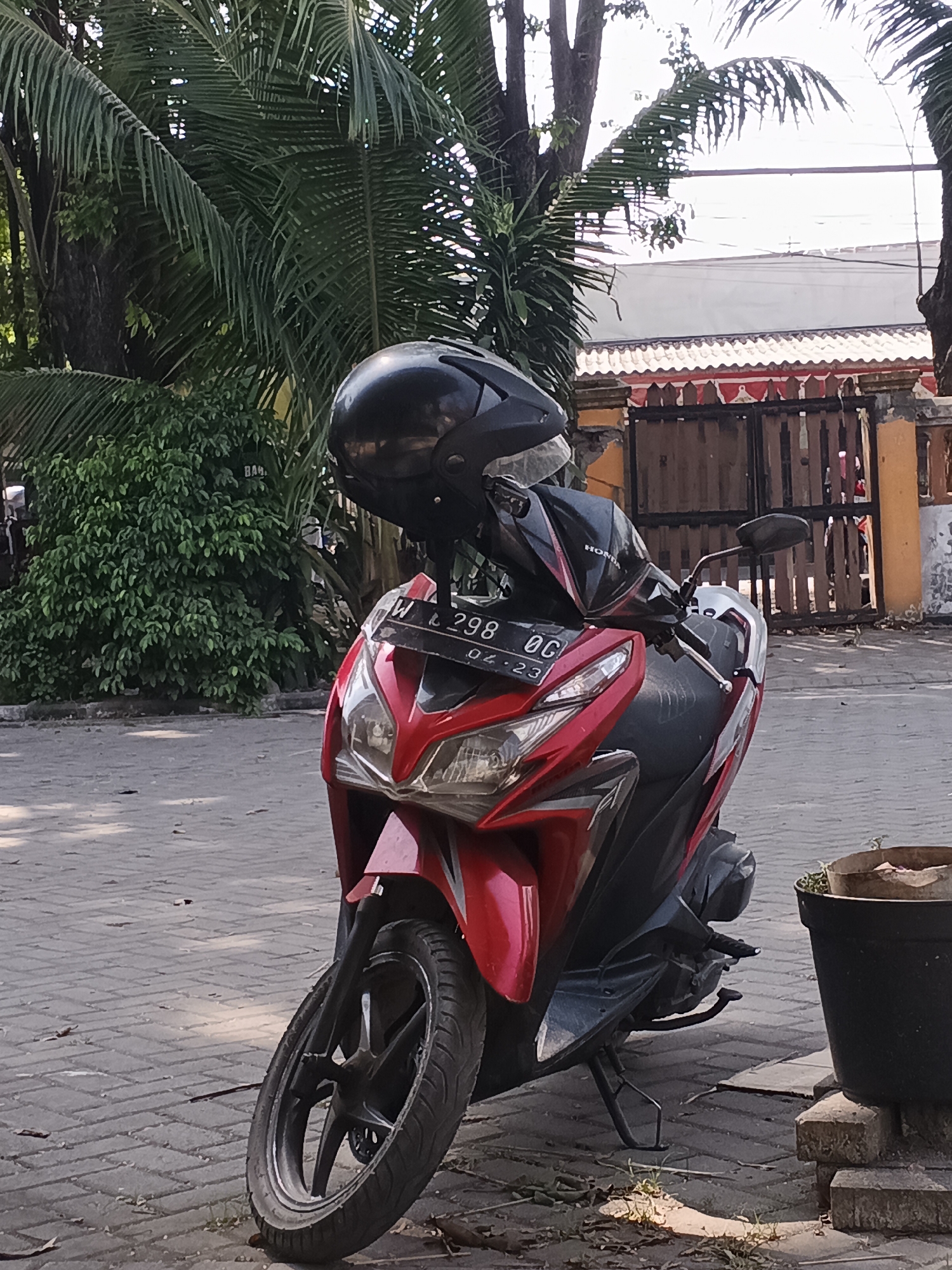 A red and black Honda scooter is parked on a paved sidewalk in Krian, Indonesia. A black helmet rests on the handlebars of the scooter. The scooter's license plate, which reads "W L 298 OG 06-23," is visible. Behind the scooter, a wooden fence with an orange and white decorative trim runs along the property line. Lush green trees and palm fronds frame the scene. The ground is paved with interlocking brick pavers, and fallen leaves are scattered across the surface. A dark pot with a small plant sits to the right of the scooter. The sky is bright and overcast, suggesting daytime.