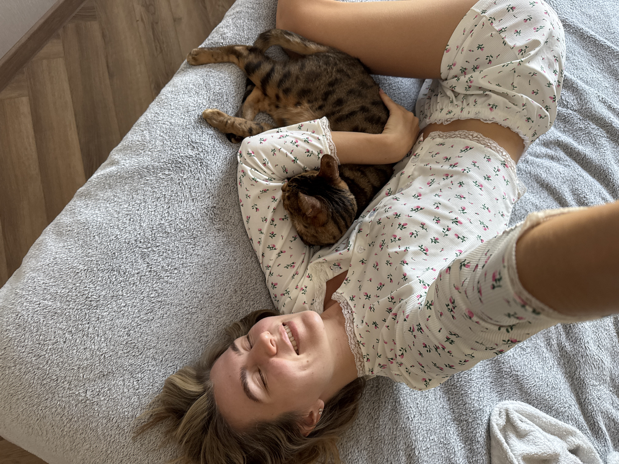 The image captures a candid moment of a young woman lying on her back on a light grey, textured blanket or comforter, presumably indoors on a bed or sofa. She has light-colored hair, a broad smile, and her eyes are gently closed, conveying a sense of happiness and relaxation. She is wearing white loungewear or pajamas adorned with a delicate pattern of small pink and green flowers.
Cuddled closely to her body and held by her arm is a domestic cat with distinctive tabby or Bengal-like markings, featuring brown and black stripes. The cat appears to be nestled comfortably against her. The woman's pose, with her arm extended upwards, suggests she is taking a selfie from an overhead perspective.
In the background to the left, a light brown wooden floor is visible, indicating an indoor setting. The lighting is bright and natural, suggesting it is daytime. A white towel or a piece of white fabric is also partially visible on the grey blanket in the lower right corner. The overall scene depicts a peaceful and affectionate interaction between the woman and her pet. There is no visible text in the image.