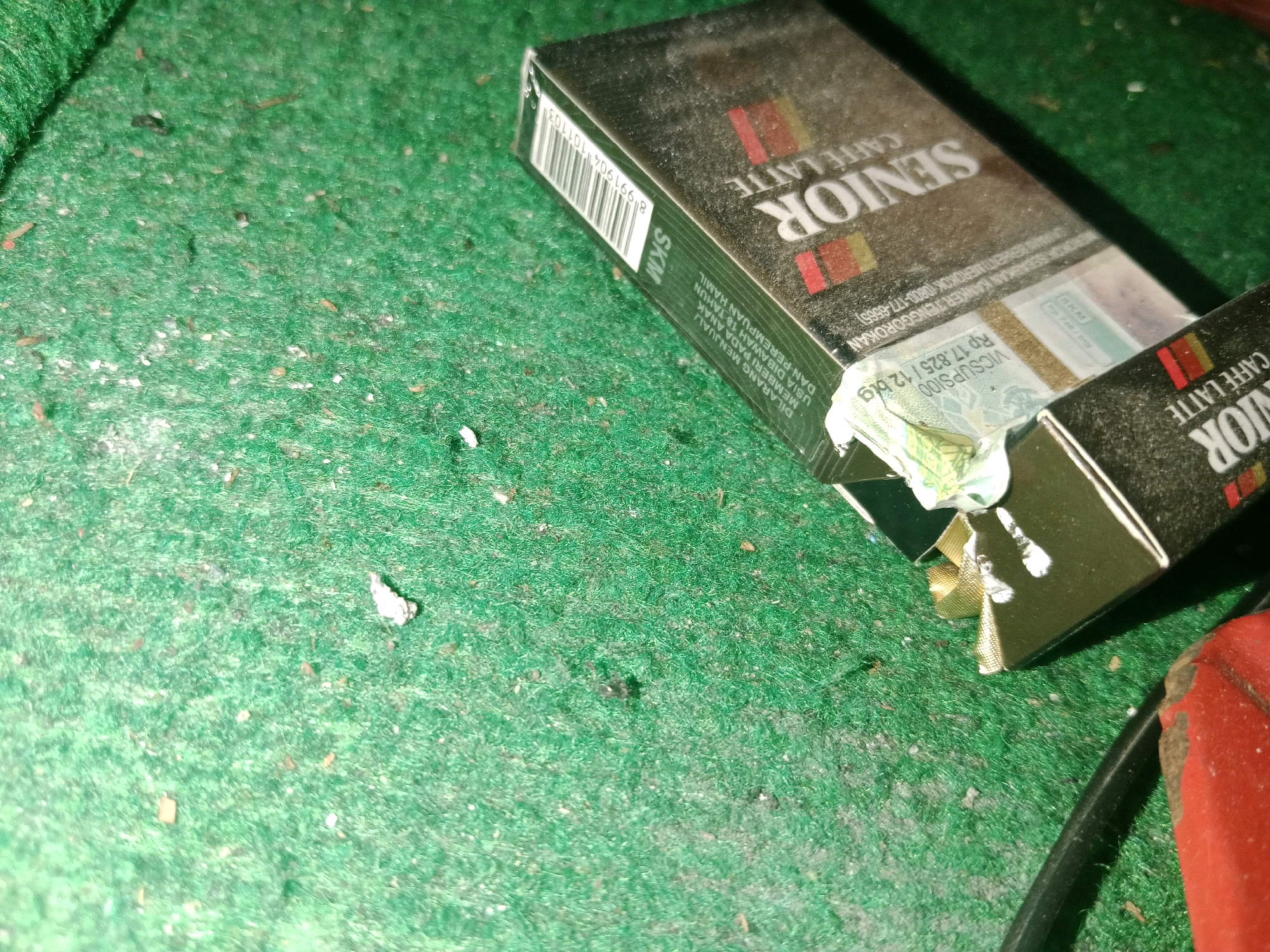 Two packs of "Senior" brand cigarettes, one with a "Caffe Latte" flavor, are placed on a green, fuzzy surface, possibly a carpet or rug. Debris, including small white particles and some organic matter, is scattered across the surface. One cigarette pack is partially open, revealing a band with the text "Rp 17.825 / 12 Btg". The scene is dimly lit, and a red object, possibly an electronic device or a portion of one, is visible in the lower right corner of the image, along with a black cable. The image appears to be taken indoors.