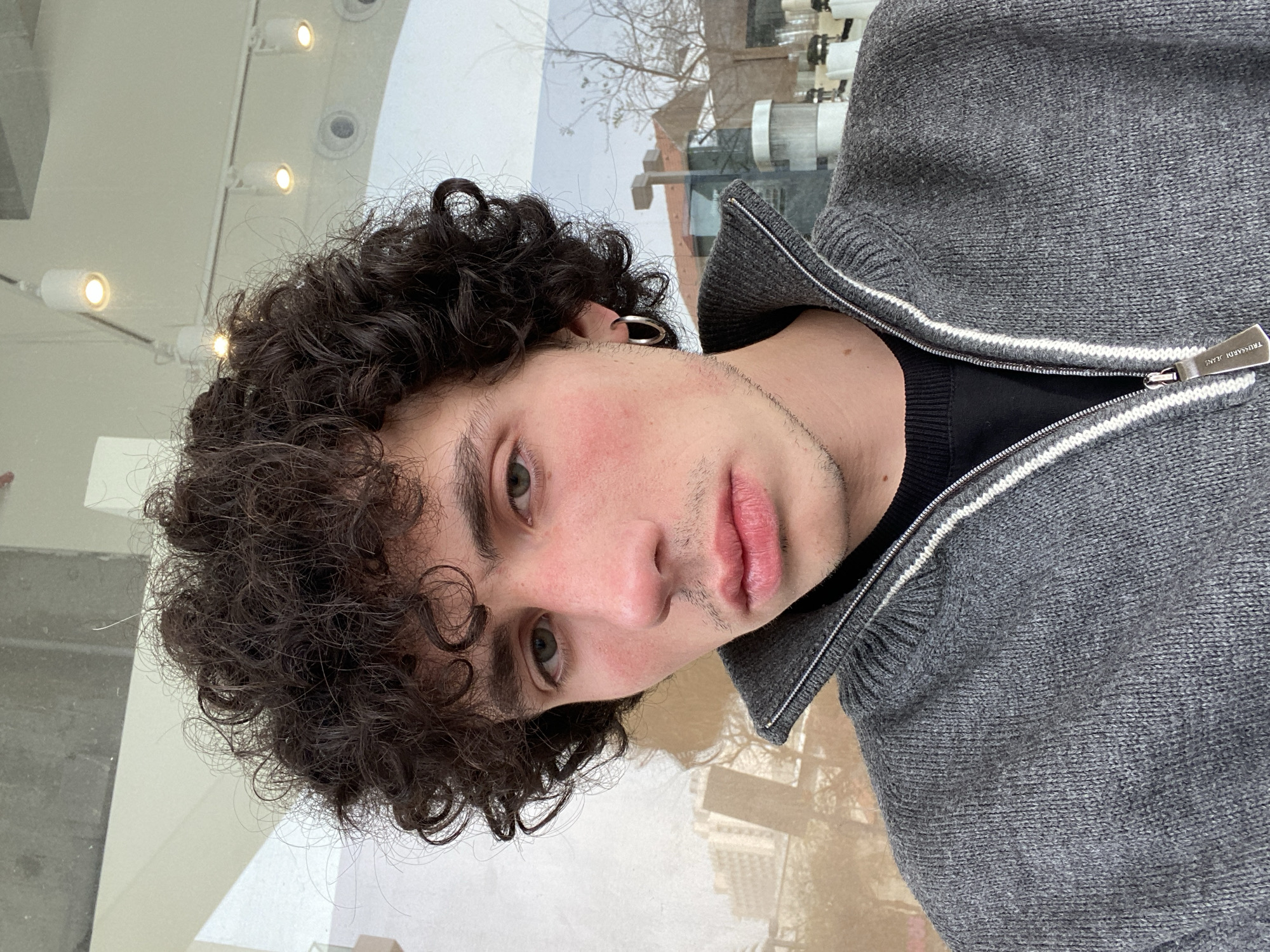 A young man with dark, curly hair and light eyes is seen in a close-up, slightly rotated selfie-style shot. He is wearing a grey knit zip-up sweater with a black undershirt. A silver earring is visible in his left ear. The background appears to be a reflection in a window. The reflection reveals a cityscape, possibly in Tel Aviv, Israel, given the architectural style of the buildings and the presence of a tree with bare branches. Indoor lighting fixtures are visible in the upper left corner, suggesting an indoor setting facing outward.
