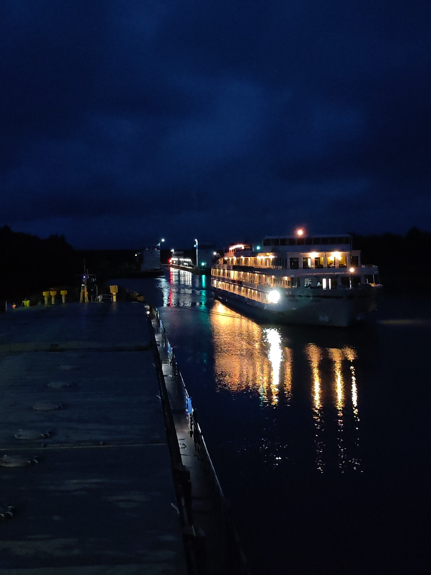 A large, illuminated passenger ship is docked on a dark body of water, likely a river or canal, at night. The ship is white with multiple decks and is aglow with interior lights, creating a warm reflection on the water. In the foreground, a portion of a dark, possibly industrial dock or barge is visible, extending into the frame from the left. The background features a dark, treeline and a distant structure with lights, suggesting it is a port or a lock system. The sky is dark blue and cloudy. The location is identified as Devyatiny, Russia. There are no people clearly visible in the image. The overall scene depicts a quiet, nocturnal maritime environment.