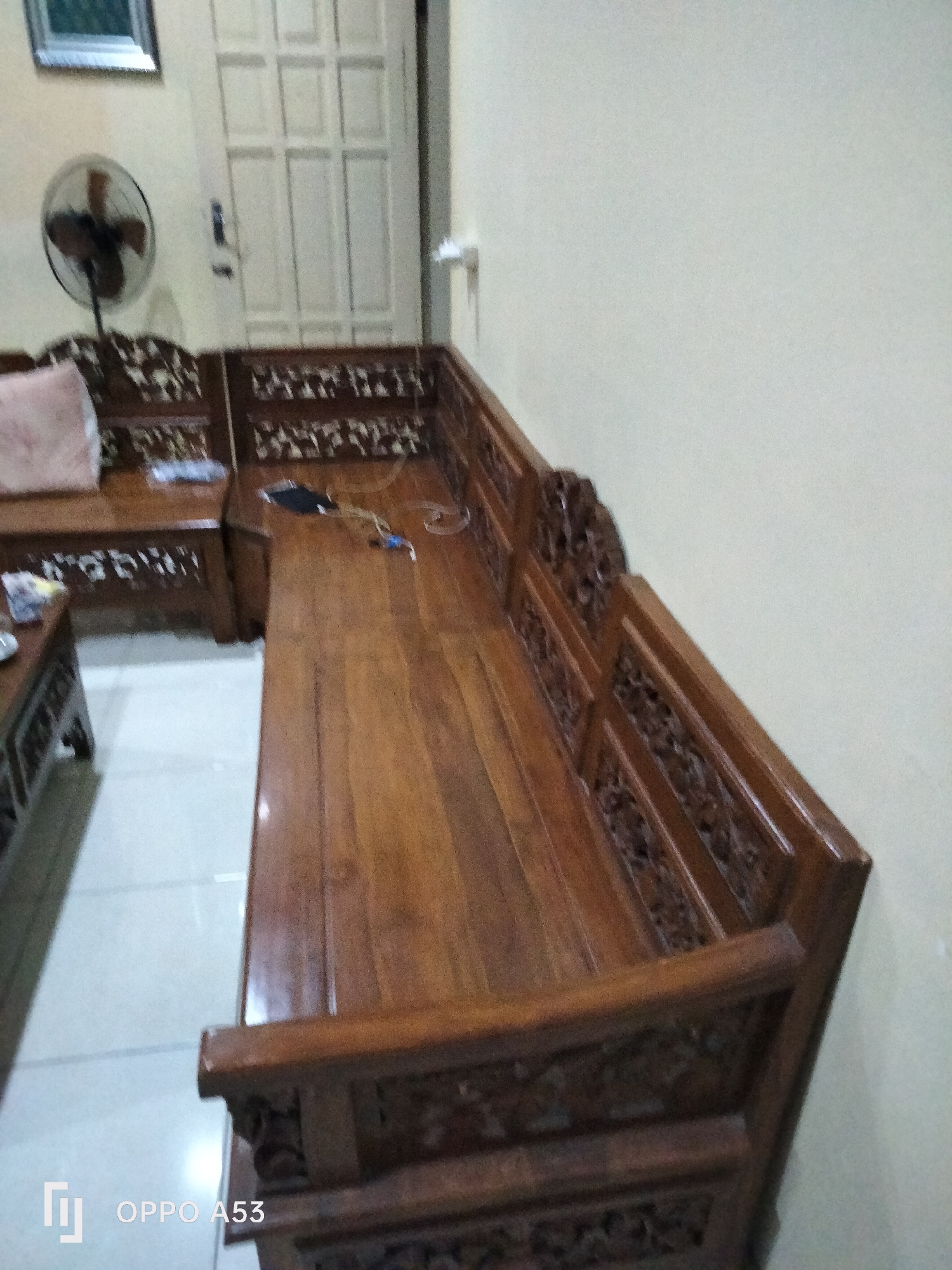 In a room, a dark wood sectional sofa with ornate carvings is positioned in a corner. The sofa has a light-colored cushion on the left section and a dark object with wires and a light on the floor to the right. The room has light-colored walls and a white door with paneling. A black pedestal fan is visible on the left, and a framed picture hangs on the wall above it. To the left of the sofa, a dark wood coffee table with a light-colored surface is partially visible. The flooring appears to be a light-colored tile. The image also displays a watermark in the bottom left corner that reads "OPPO A53." The setting is the city of Rejowinangun, Indonesia.