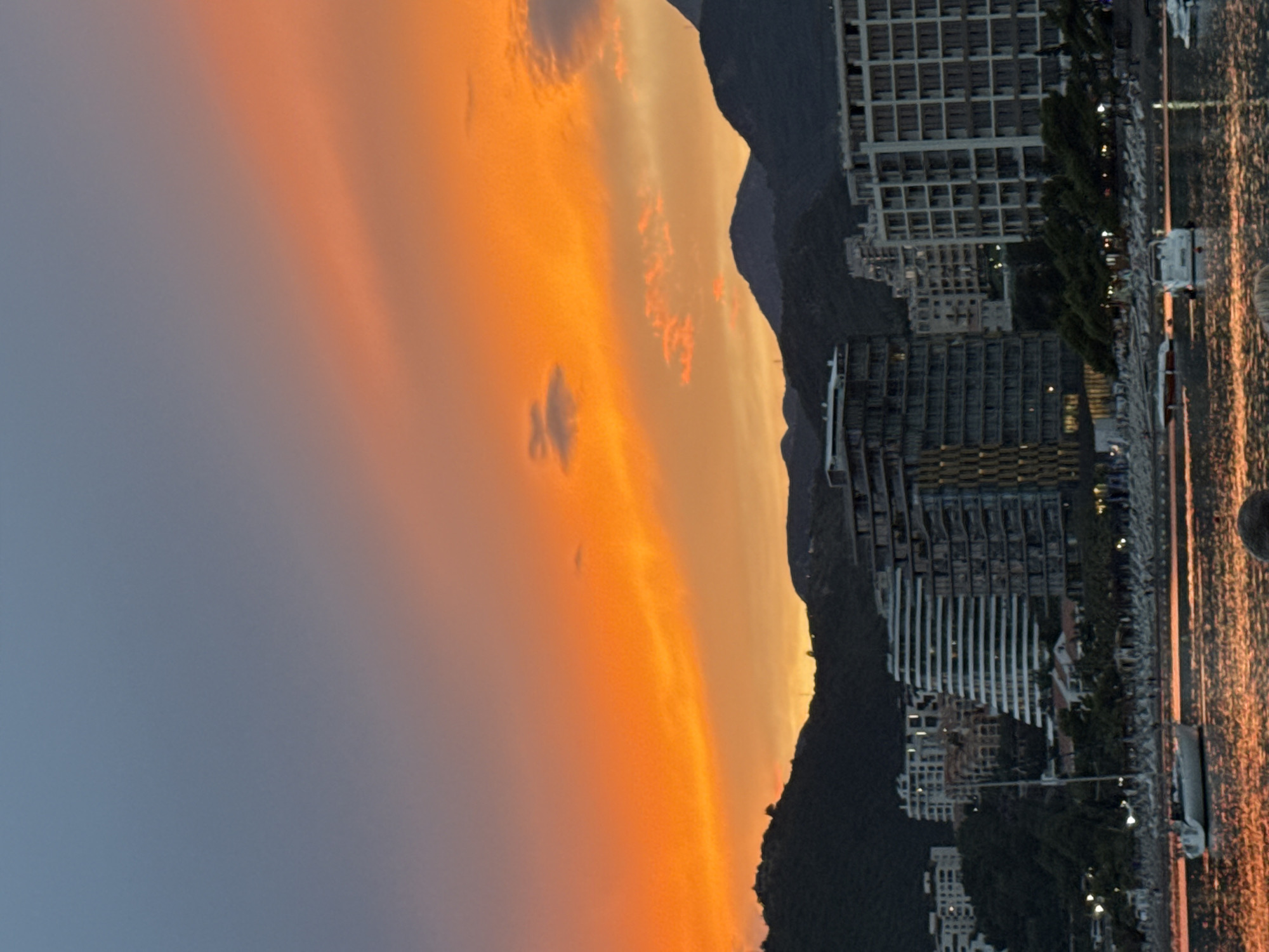 This is a nighttime photograph of a coastal town with a dramatic sky. The sky is filled with vibrant orange and yellow hues, suggesting a sunset or sunrise, with streaks of darker clouds. In the foreground, there is a body of water reflecting the colors of the sky, creating a shimmering effect. The water appears calm. Along the shore, there is a row of buildings, some of which are illuminated, indicating that it is likely evening or early morning. These buildings are modern in design, with multiple stories and many windows. Trees are also visible along the waterfront. The scene is bathed in the warm glow of the sky and artificial lights. There are no people or specific objects clearly identifiable as the main subject, but the overall impression is one of a peaceful, scenic waterfront at dusk or dawn. The location is Rafailovići, Montenegro.