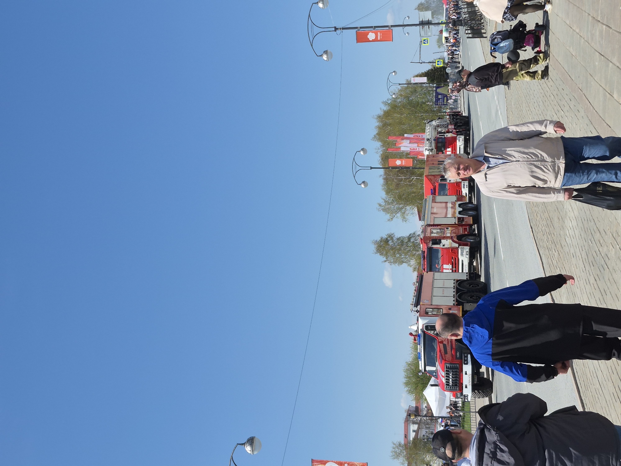 The image shows a street scene in Tobolsk, Russia. Several fire trucks are parked along the side of the road. A crowd of people is visible in the background, suggesting an event or parade is taking place. Several individuals are walking along the sidewalk; one person is wearing a blue jacket and another person is wearing a tan jacket. Overhead, lampposts are spaced along the street, and red banners hang from them. The sky is clear and blue.
