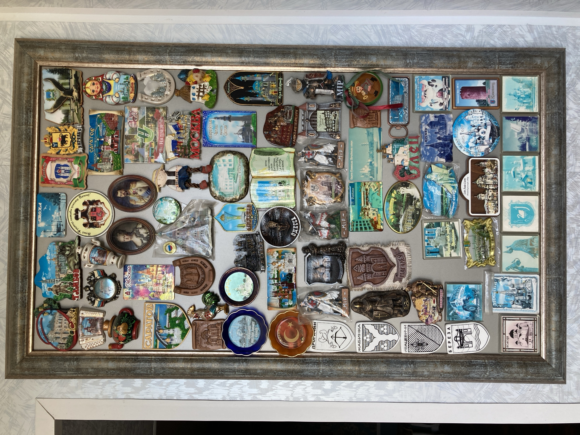 This is a close-up, eye-level shot of a framed collection of refrigerator magnets. The magnets depict various landmarks, cultural items, and scenes, primarily from Russia and Belarus, suggesting a travel-related collection. They are arranged in a grid-like pattern within a distressed, grey wooden frame.
The magnets themselves are diverse in shape and material. Many are rectangular and feature colorful illustrations or photographs of famous buildings like the Kremlin or mosques, as well as scenes of cities and natural landscapes. Others are shaped like traditional Russian nesting dolls, horseshoes, or ornate crests. Some magnets appear to be made of metal with embossed designs, while others are made of a more flexible material with printed images.
Visible text on the magnets includes place names such as "Москва" (Moscow), "Санкт-Петербург" (Saint Petersburg), "Казань" (Kazan), "Саратов" (Saratov), "Одесса" (Odessa), and "ЖАБИНКА" (Zhabinka), as well as phrases like "Я хочу Вам пожелать" (I want to wish you) and "Belle Russe" (White Russia). The overall impression is a personal collection showcasing memories and experiences from various travels. The lighting is neutral, suggesting an indoor setting, and there are no indications of time of day or weather. The composition is straightforward, focusing entirely on the framed collection.