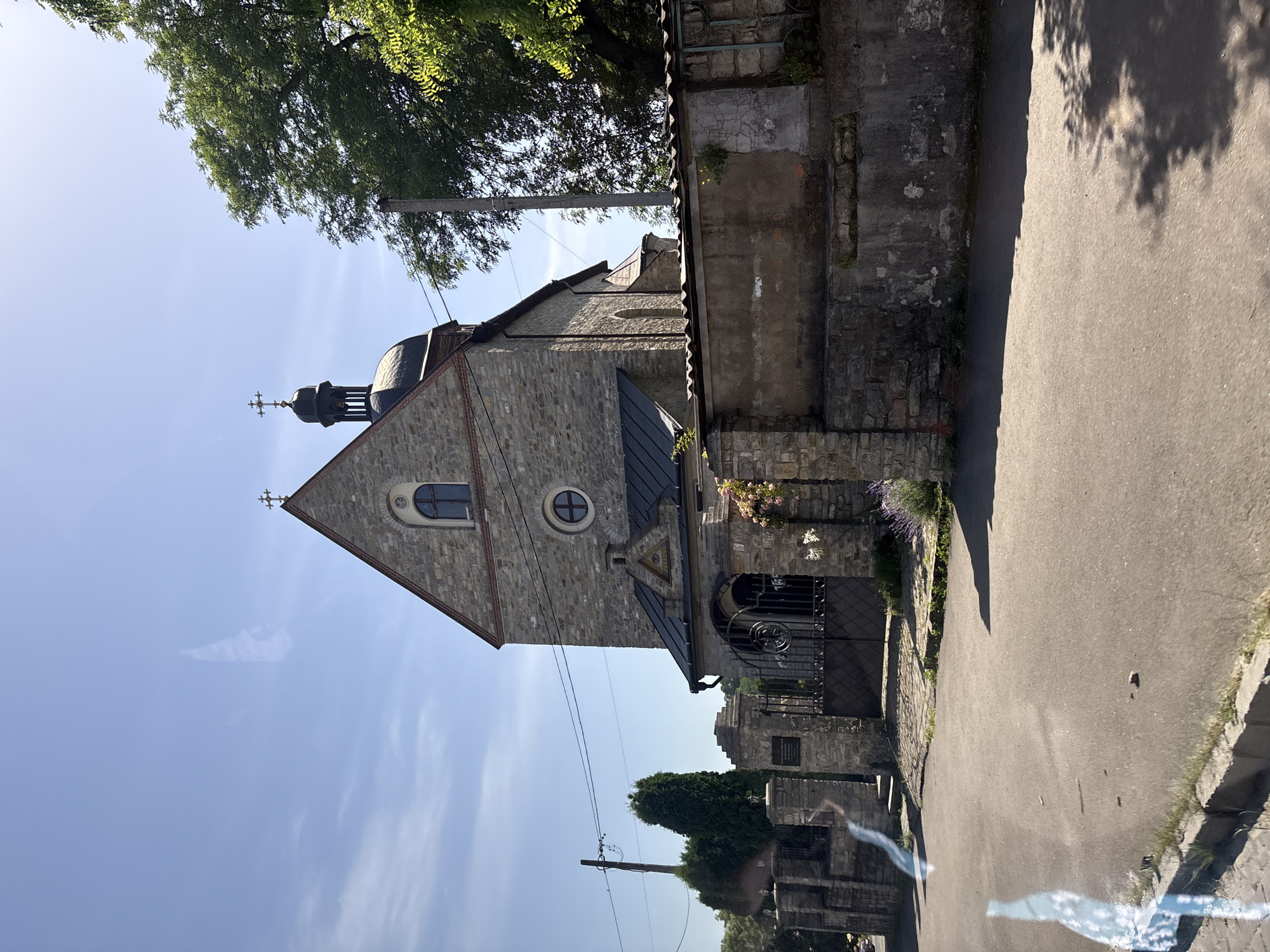 A stone church with a dark dome and a cross on top stands under a clear blue sky. The building is partially obscured by a stone wall and fence. A wrought iron gate leads to the church entrance. Trees are visible on the right side of the image. The scene appears to be in a rural or suburban setting.
