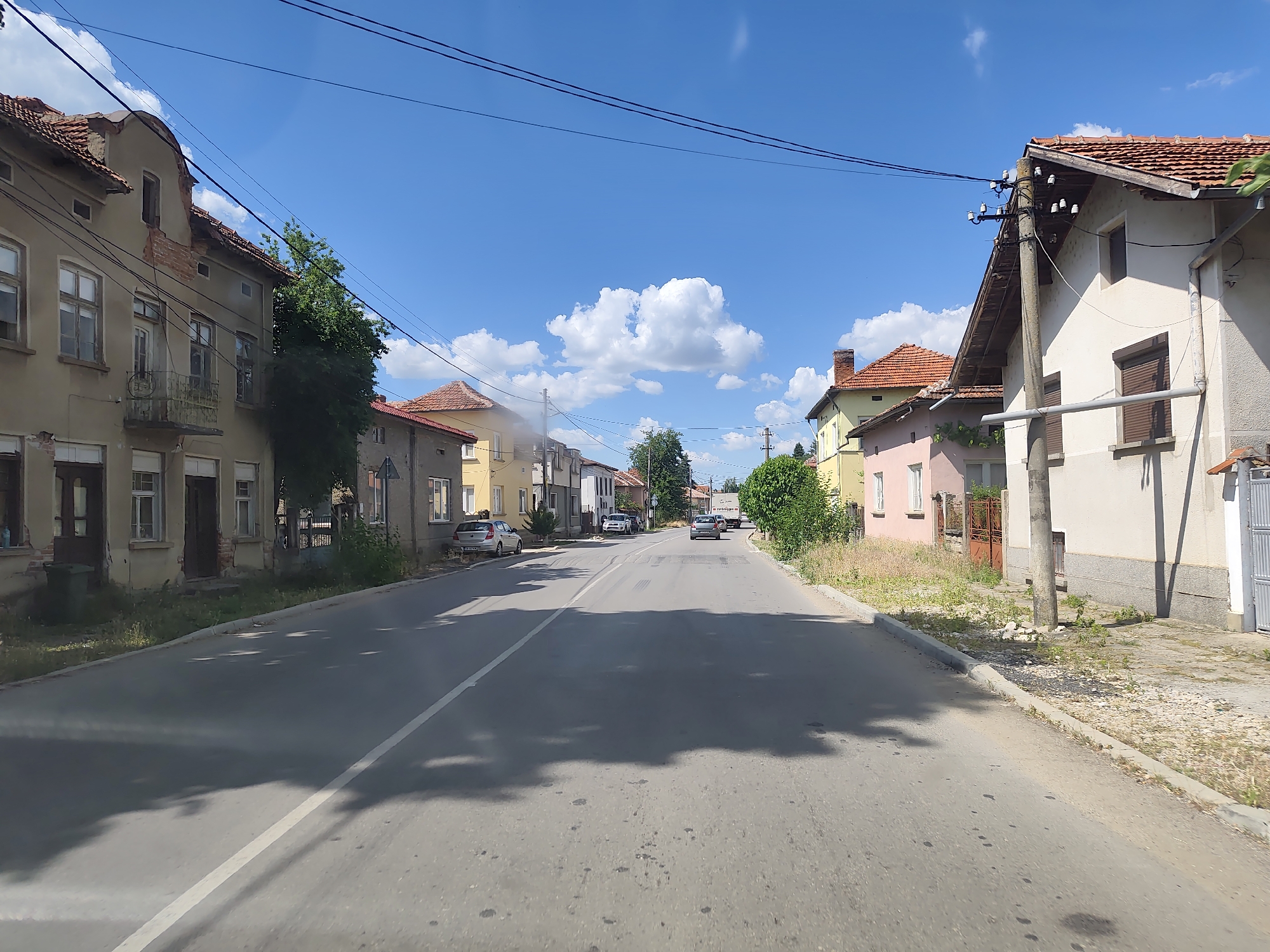 The image depicts a street scene in Dermantsi, Bulgaria, on a sunny day. The sky is bright blue with scattered white clouds, indicating fair weather. The time of day appears to be midday or early afternoon due to the strong sunlight and distinct shadows cast by trees and overhead wires.
The scene is a relatively narrow, paved road lined with residential buildings on both sides. The architecture of the houses is varied, with some appearing older and somewhat dilapidated, showing exposed brickwork and peeling paint, while others are more modern and well-maintained. Red tiled roofs are a common feature across many of the buildings.
Several cars are parked along the street or are in motion. On the left side of the road, there's an older, multi-story building with balconies and visible windows. Adjacent to it, further down the street, are other houses and trees. On the right side, a more modern-looking house with a cream-colored facade and brown shutters is prominent, with a utility pole situated in front of it.
The road itself has a white line marking the center and appears to be in moderate condition with some patches and markings. Shadows of trees are cast across the asphalt, suggesting the presence of foliage along the roadside. The overall impression is of a quiet, lived-in village or small town environment. No people are clearly visible in the image. There is no discernible text in the image.