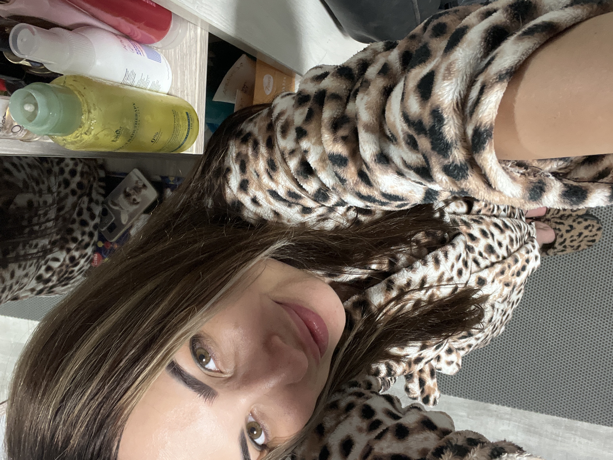 An adult female is depicted in a high-angle selfie. She has long, straight brown hair with lighter highlights, brown eyes, and well-groomed eyebrows. Her lips are subtly glossed, and she exhibits a faint smile. She is wearing a plush robe with a brown, black, and cream leopard print pattern. Matching leopard print slippers are visible on her feet on the left side of the frame. The setting is indoors, with a light gray, textured mat visible on a lighter floor in the lower left quadrant. In the upper right background, a shelf unit contains several personal care products: a prominent yellow-green bottle labeled "Lador PERFECT HAIR THERAPY" (150 ml), a white spray bottle, and a red/pink tube. A white phone case with an animal design is visible on a surface in the upper left background. The location is an interior space in Unknown, Unknown.